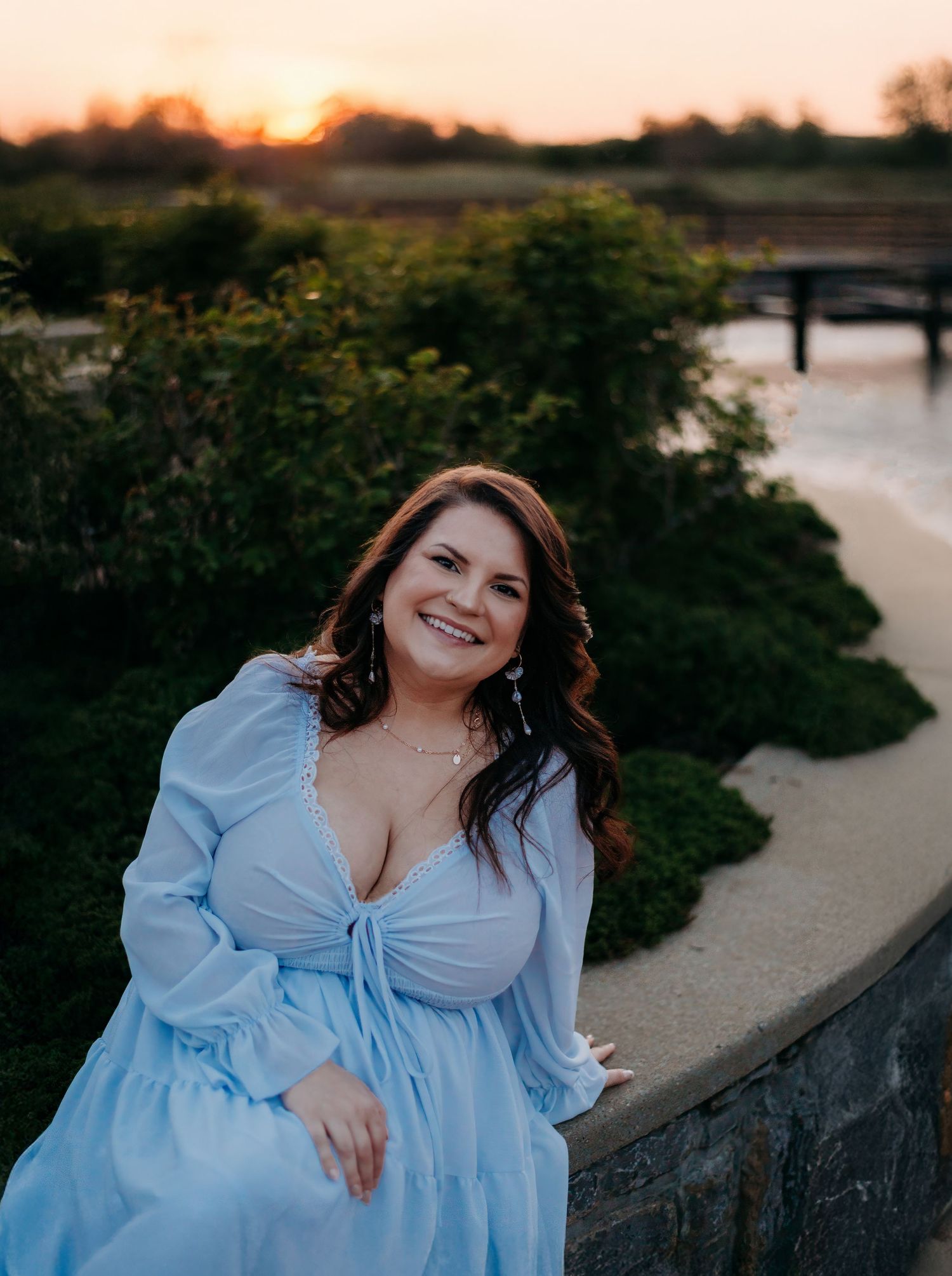 A woman in a flowing blue dress poses by a waterfront at sunset.