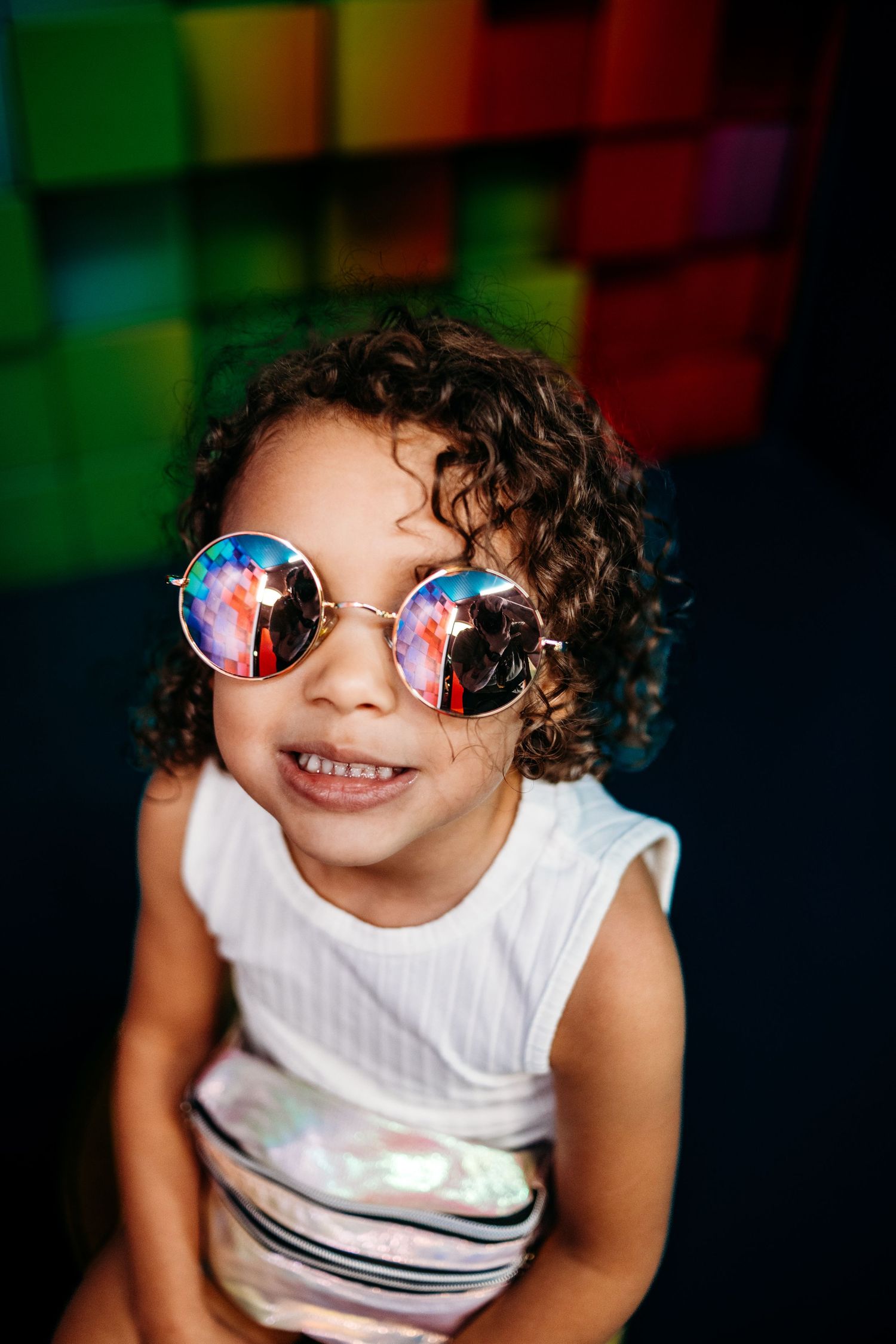 A child wearing round reflective sunglasses smiles against a colorful geometric background.
