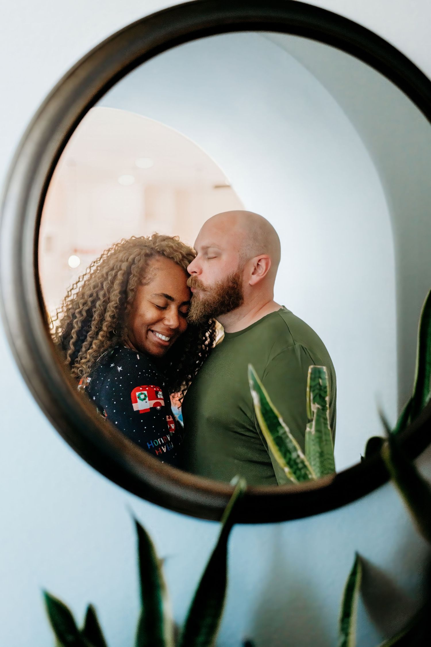 A couple shares a tender moment reflected in a round mirror with plants visible.
