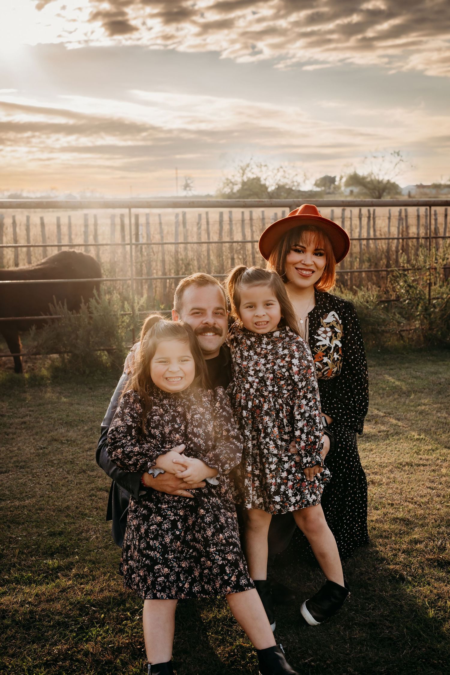 Family group wearing floral dresses sitting together in field at sunset.