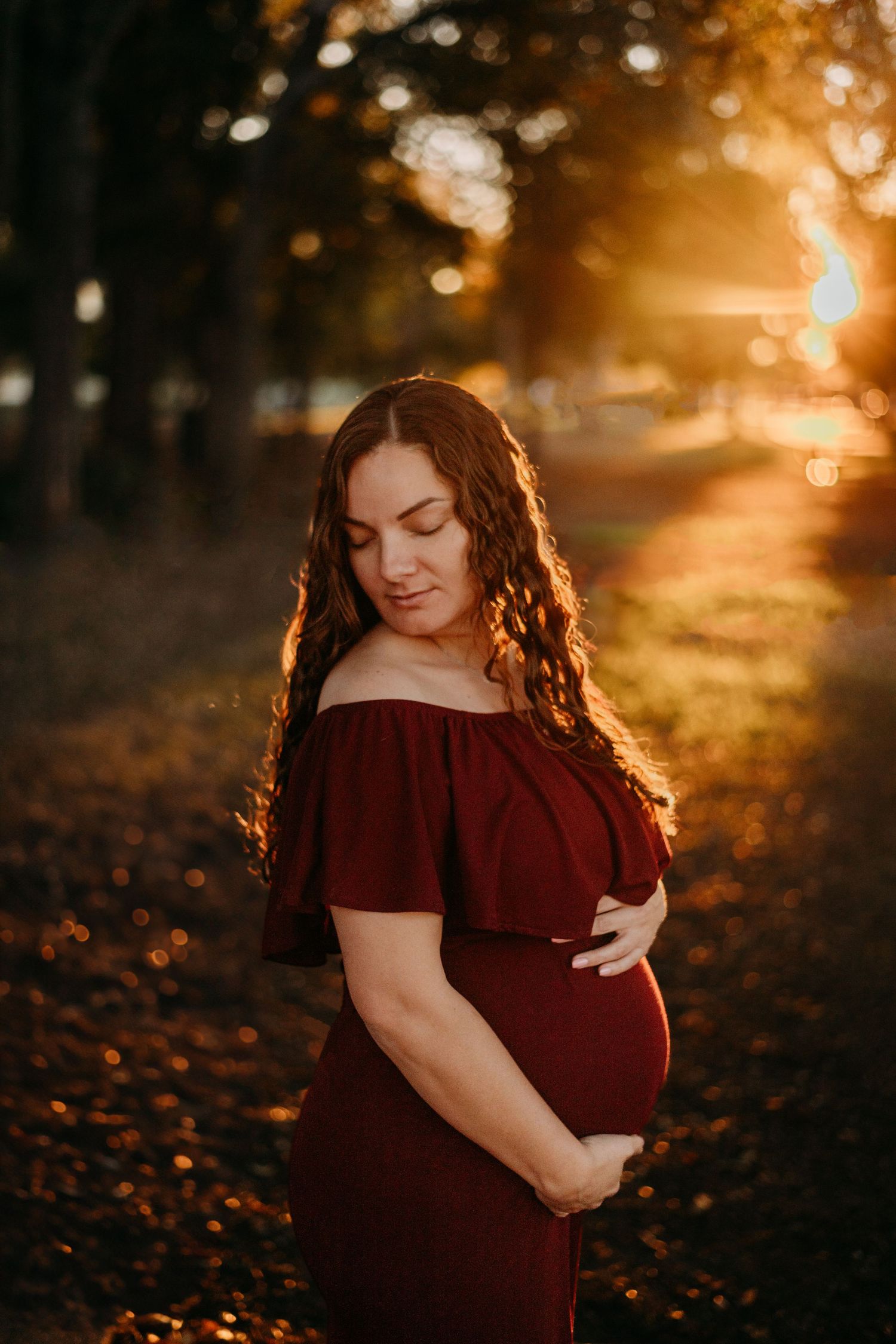 An expectant mother in a burgundy dress poses in autumn sunlight among fallen leaves.