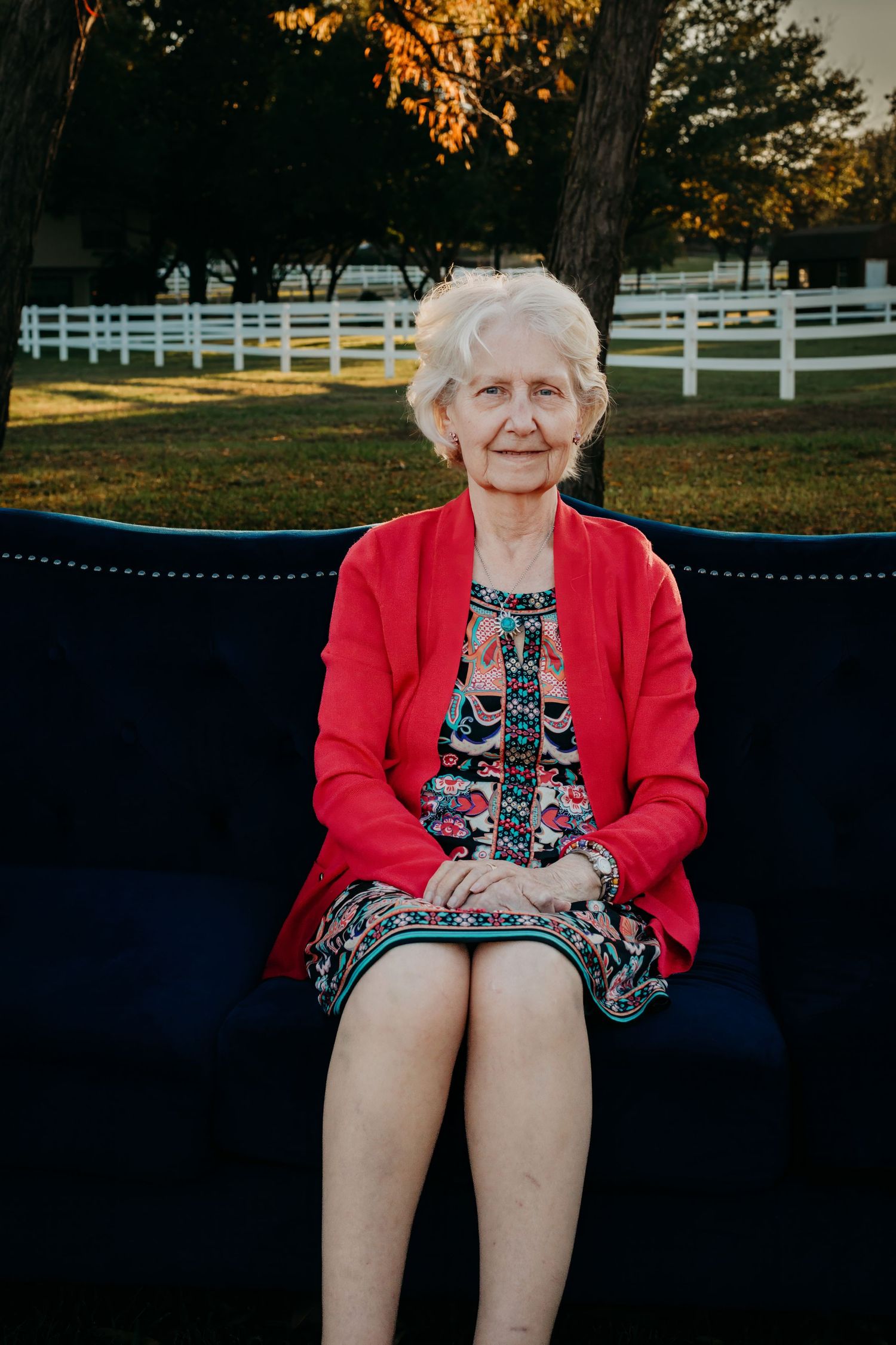 An elderly woman in a red cardigan sits on a bench with white fencing in the background.