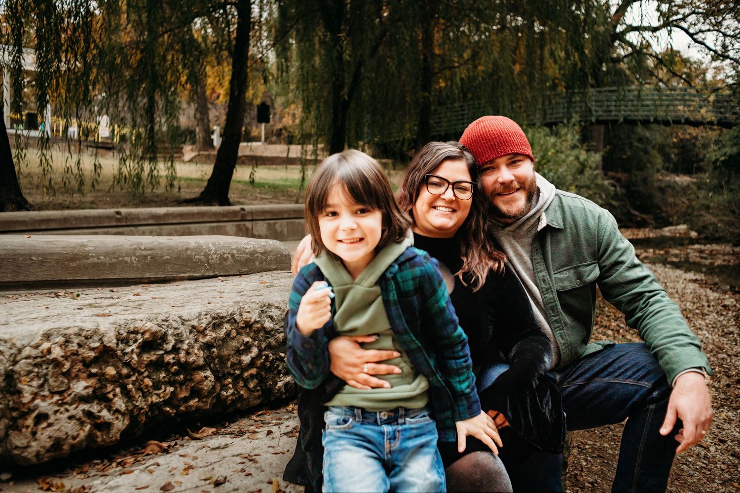 A happy family poses together outdoors on a fall day near a willow tree.