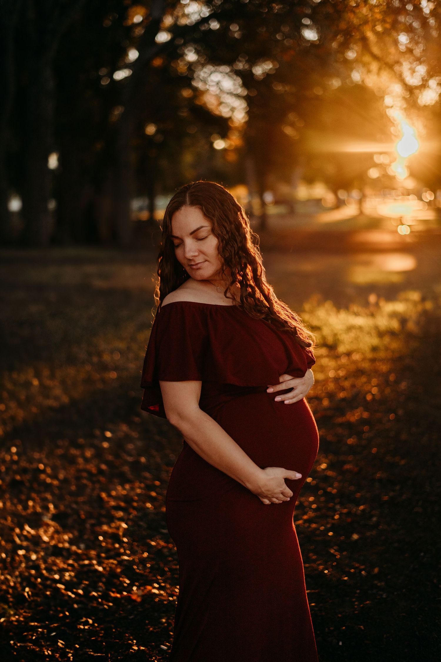 A pregnant woman in a red dress stands among fall foliage at sunset.