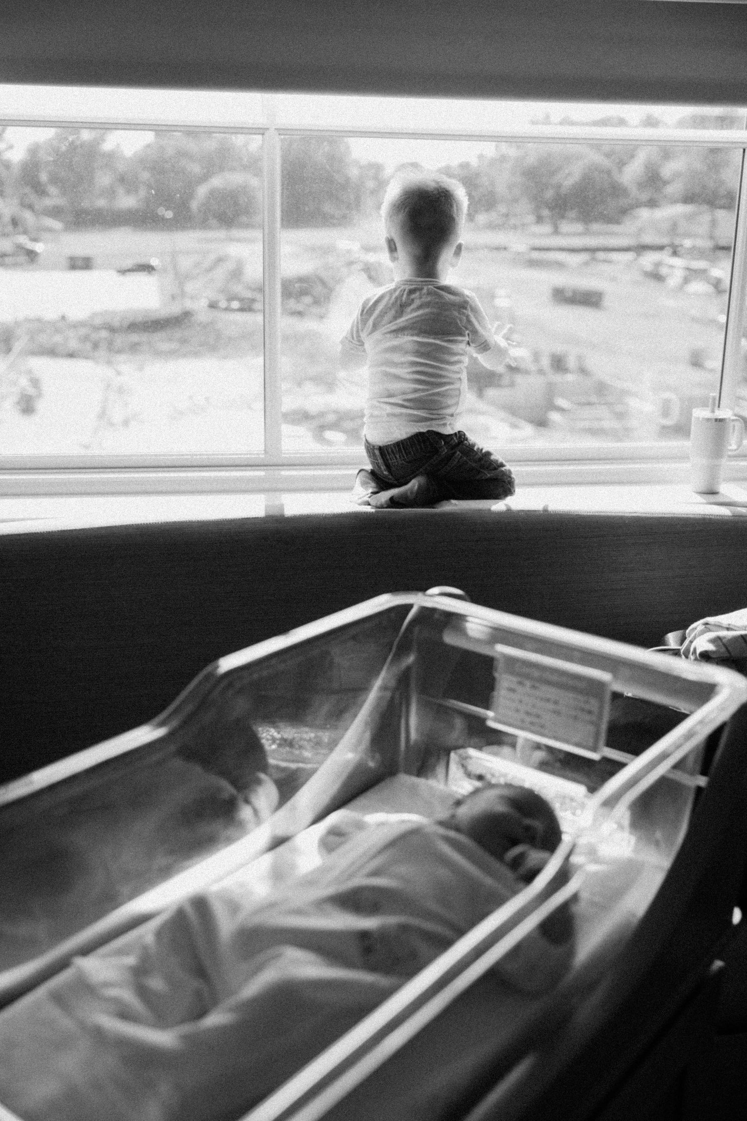Black and white photo of child looking through window with bassinet in foreground.