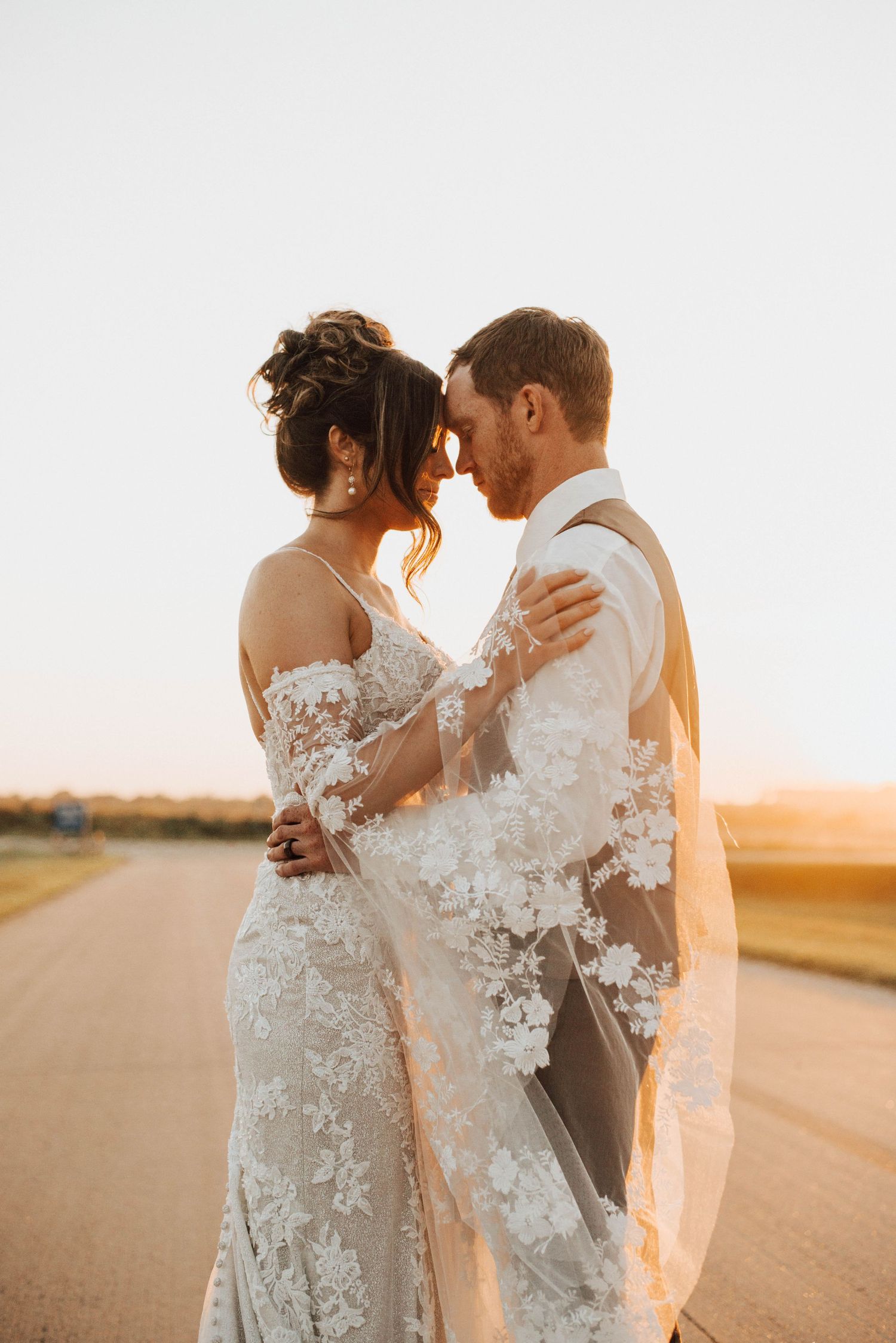 Newlyweds in lace wedding attire embracing during golden hour sunset.