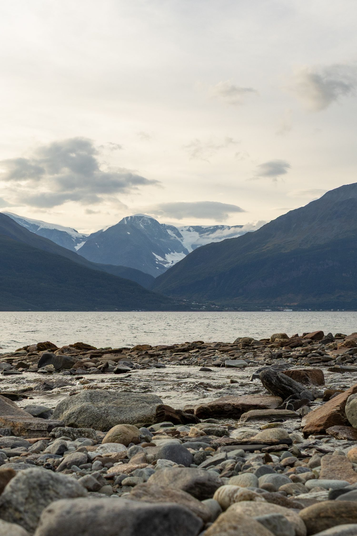 Rocky shoreline with snow-capped mountains and cloudy skies in the background.