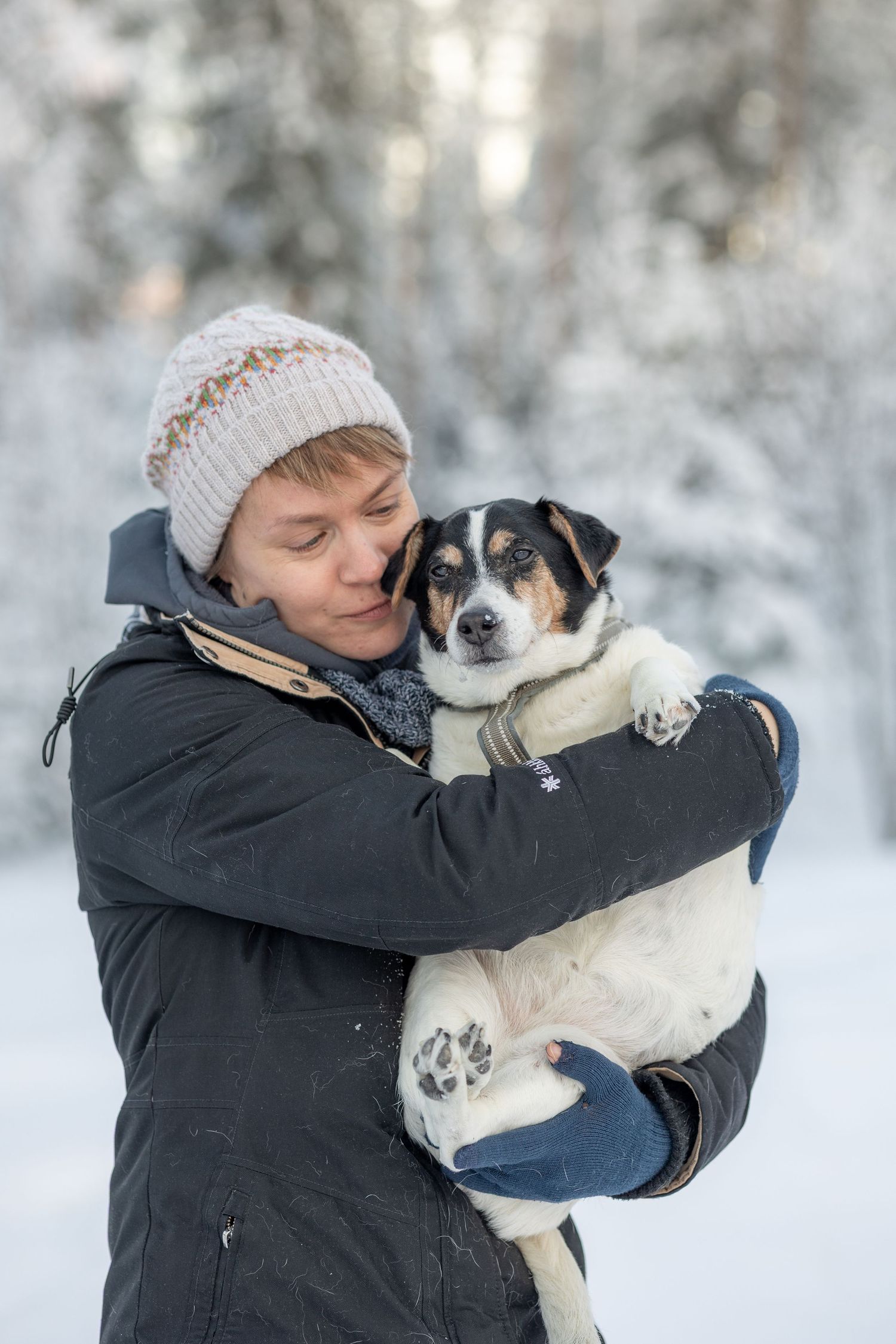 Henkilö halaa pientä kirjavaa koiraa sylissään, taustana talvimaisema.