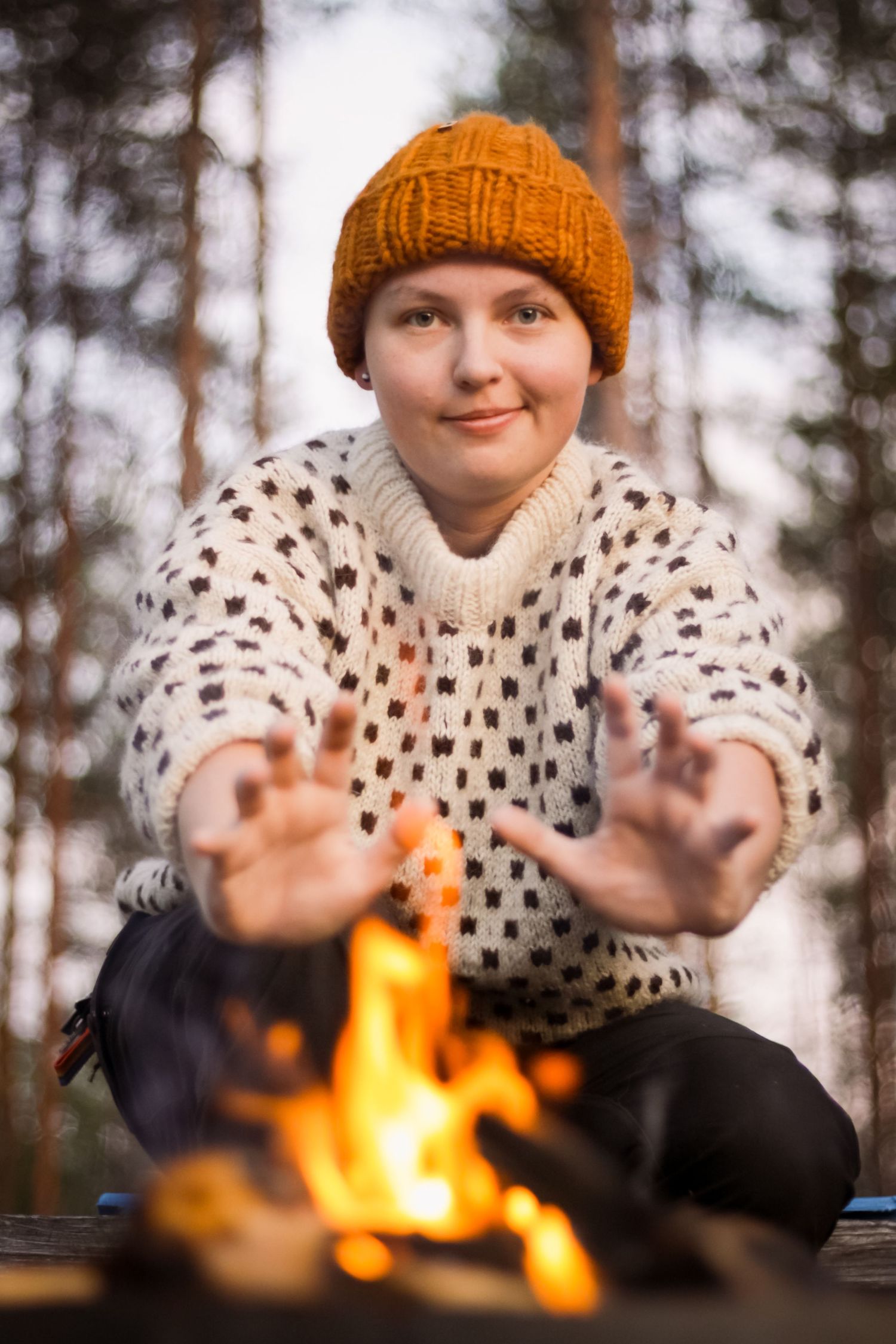 Person in knit sweater and orange beanie warming hands by campfire.