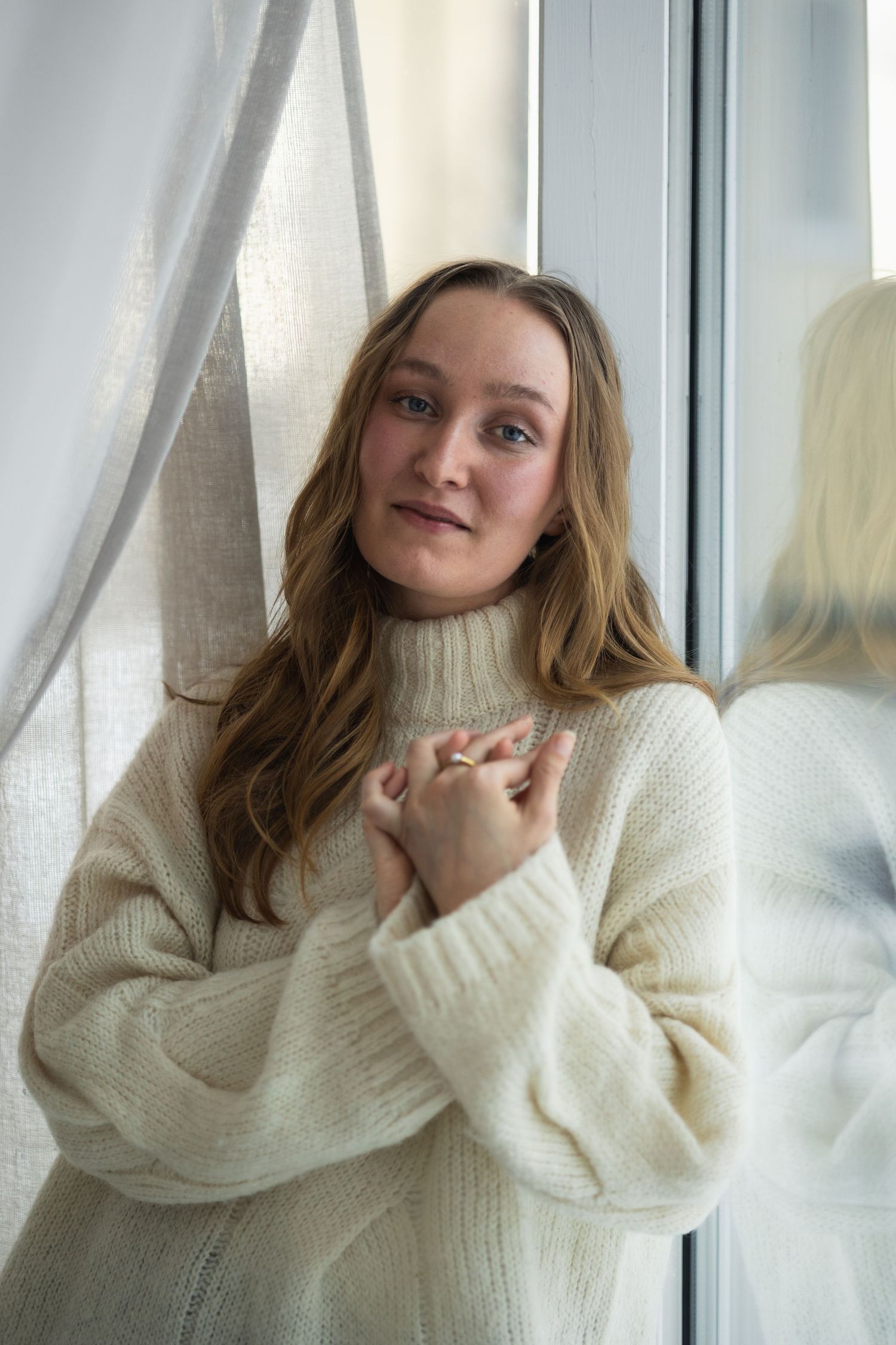 Person wearing cozy cream turtleneck sweater poses playfully near window in soft natural light.