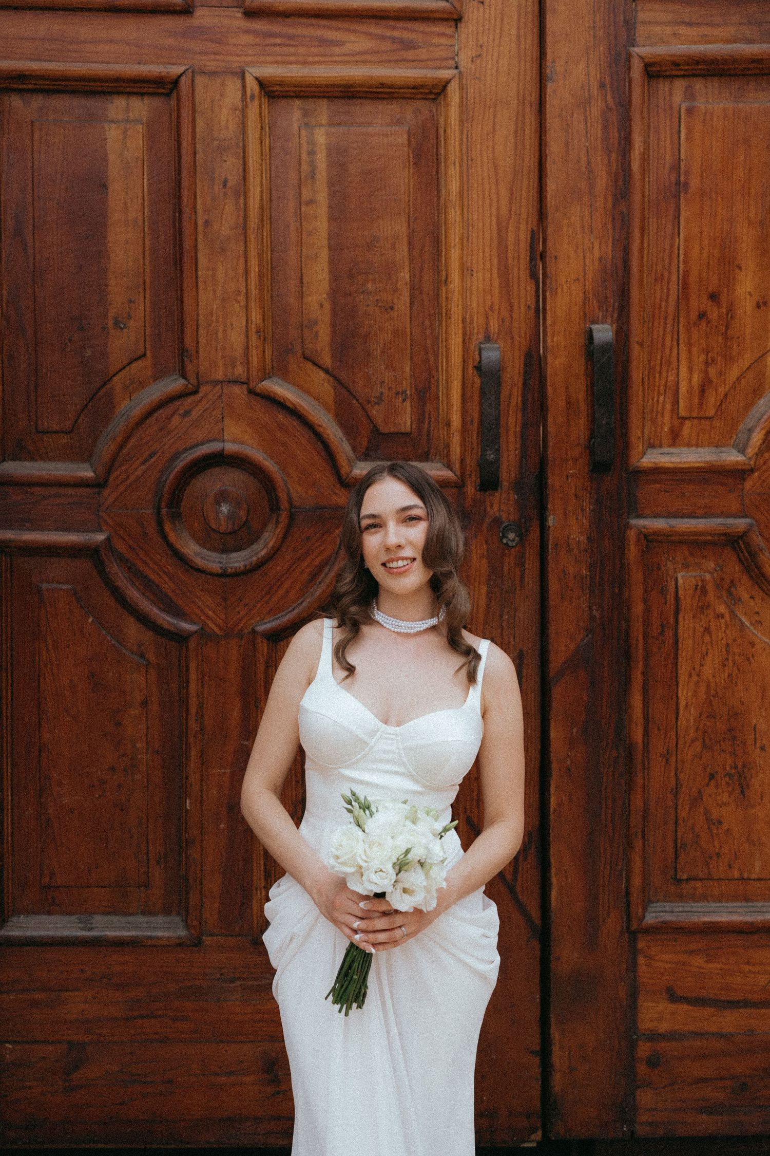 A bride in a white dress poses in front of ornate wooden doors.