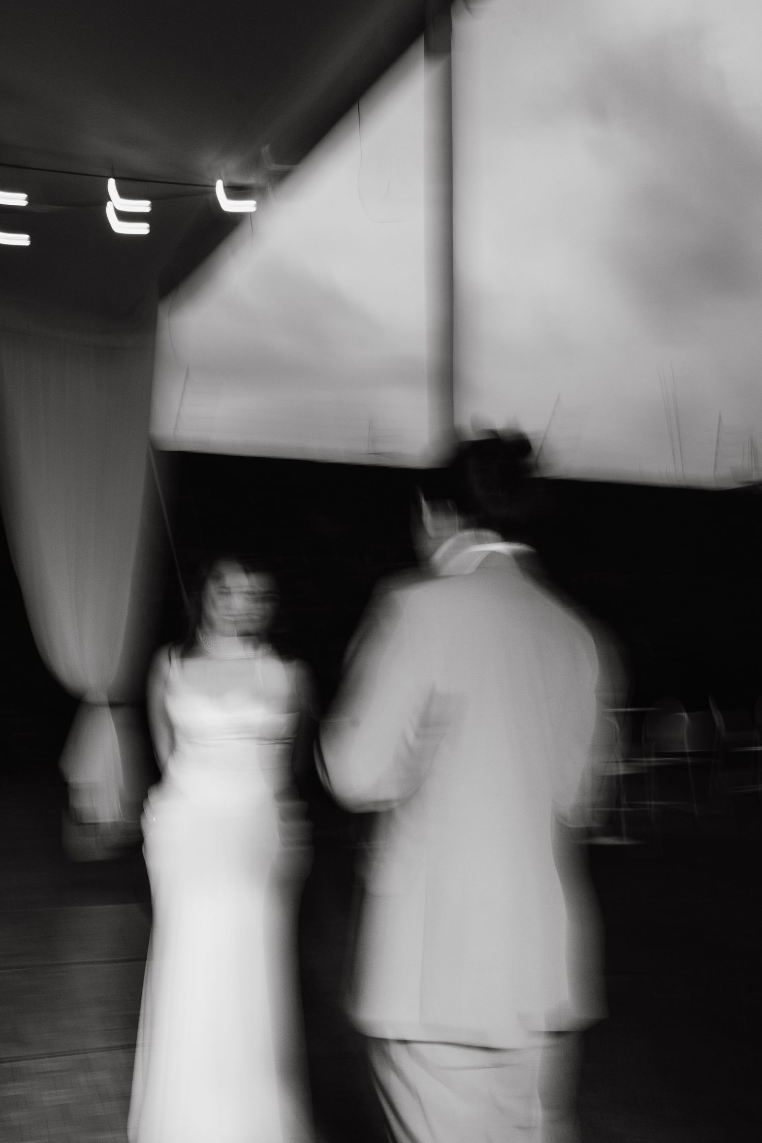 Black and white motion blur photo of dancing figures at a wedding reception under dim lighting.