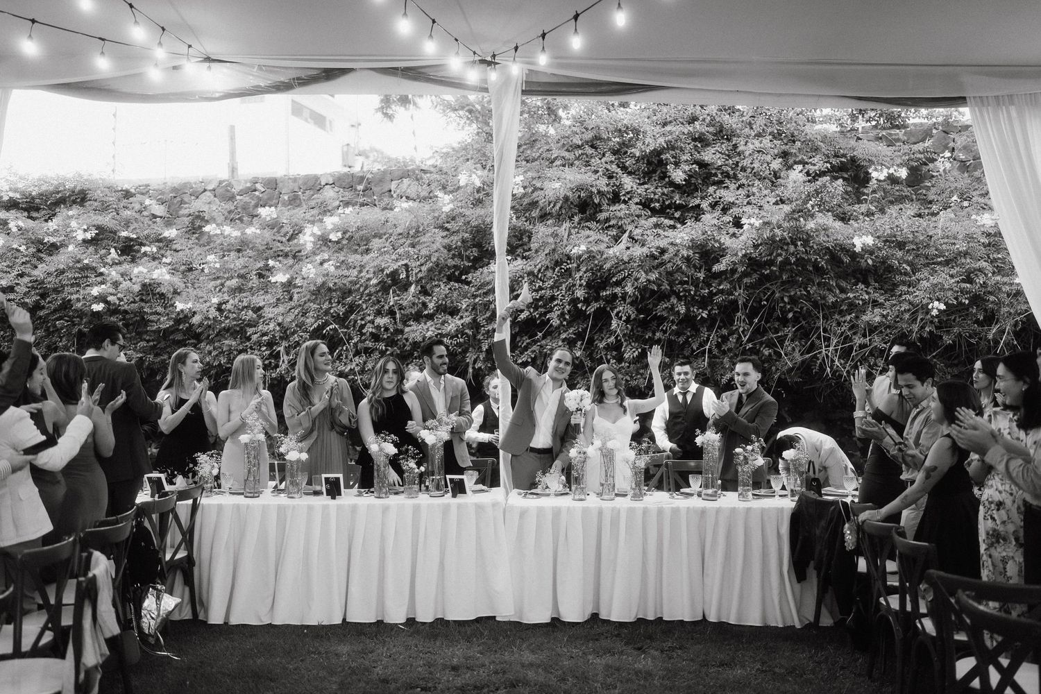 Wedding guests gather under a tent decorated with string lights during an outdoor reception celebration.