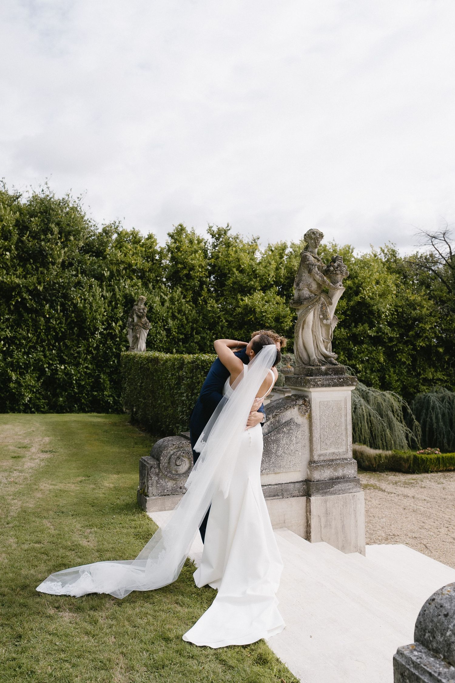 A bride in a flowing white gown poses near classical garden statues on manicured grounds.