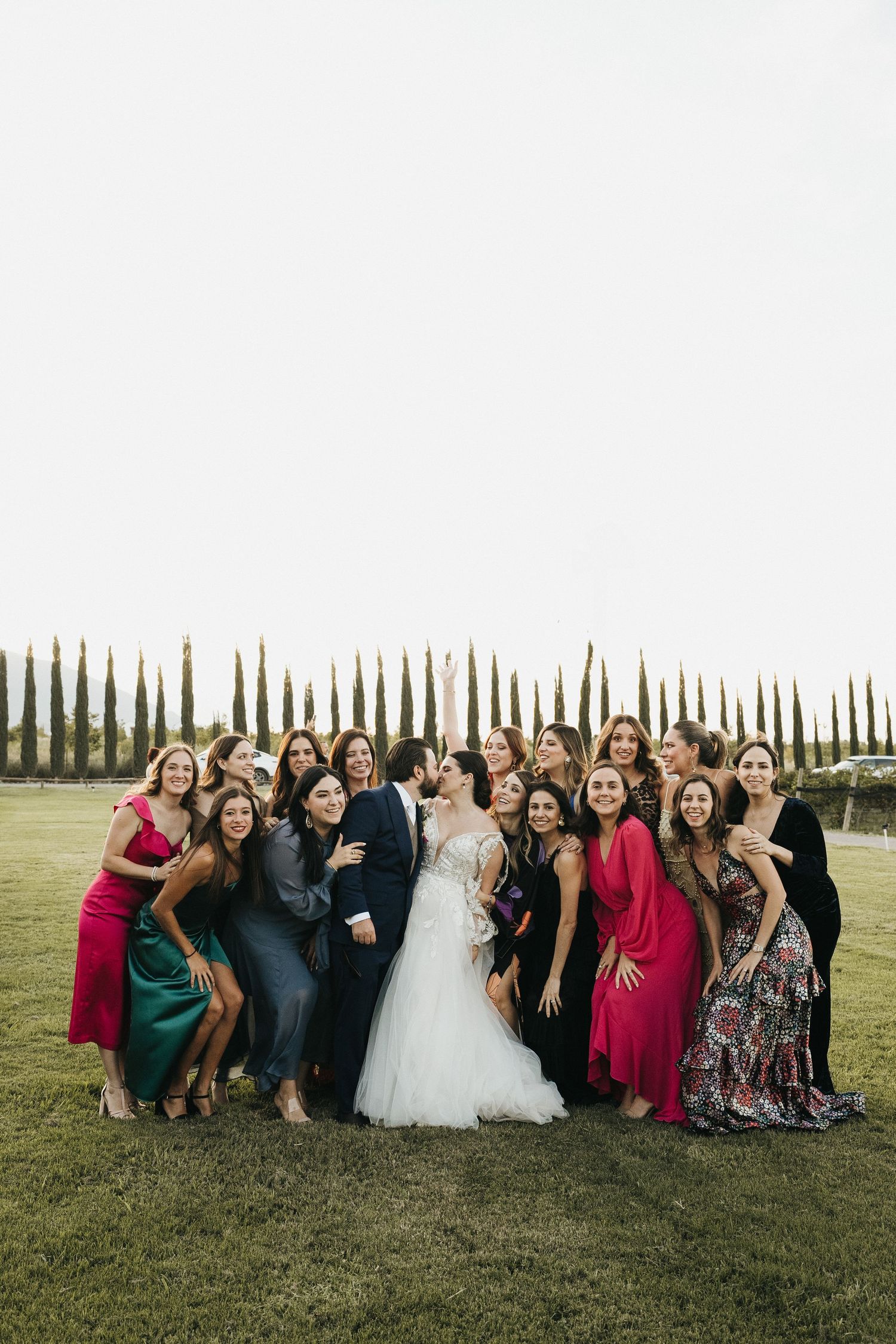 A wedding party group photo on a lawn with guests wearing colorful dresses.