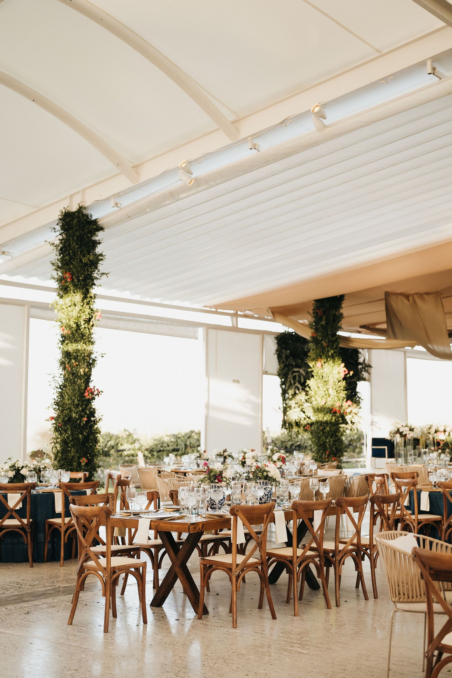 Elegant wedding reception venue with wooden cross-back chairs and tall floral centerpieces under white tent.