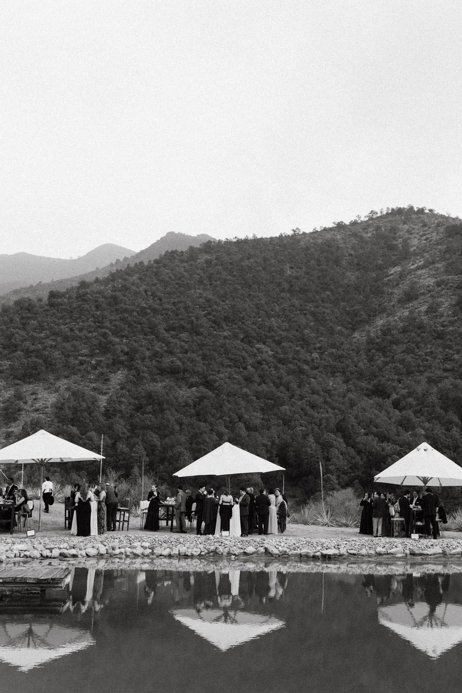 Black and white photo of wedding tents reflected in a lake with mountains in background.