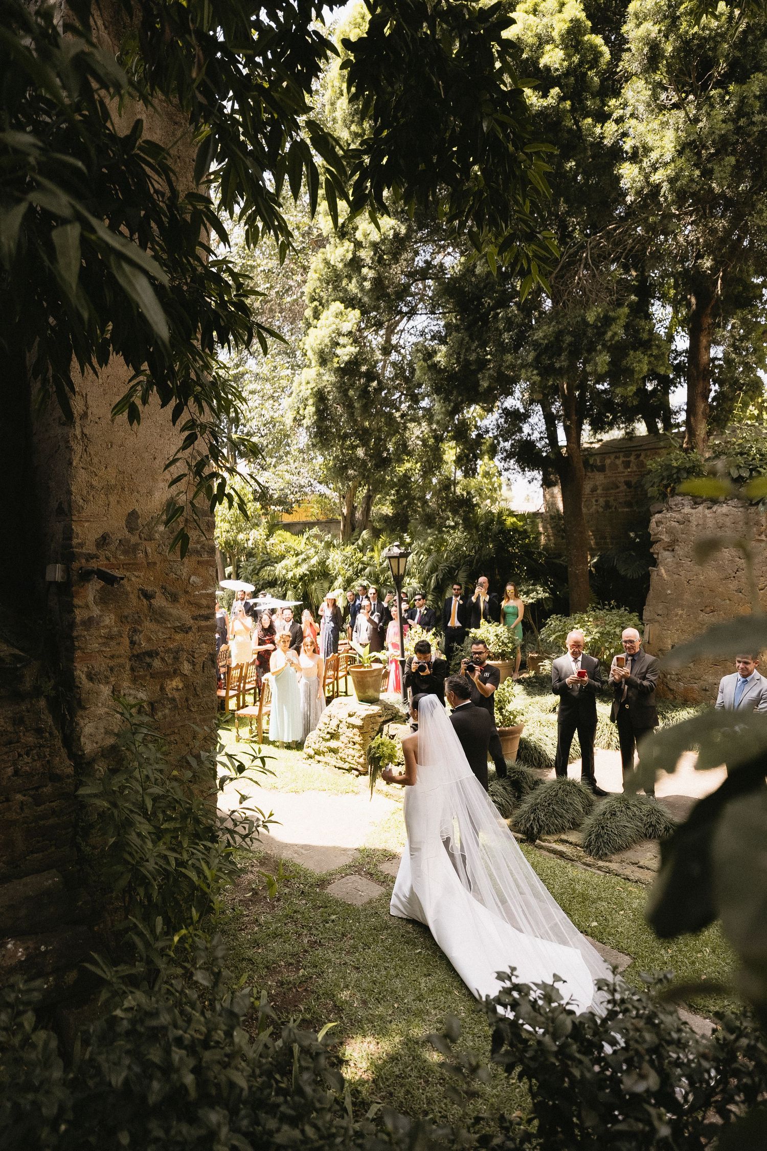 Garden wedding ceremony with guests lining stone pathways amidst lush greenery.