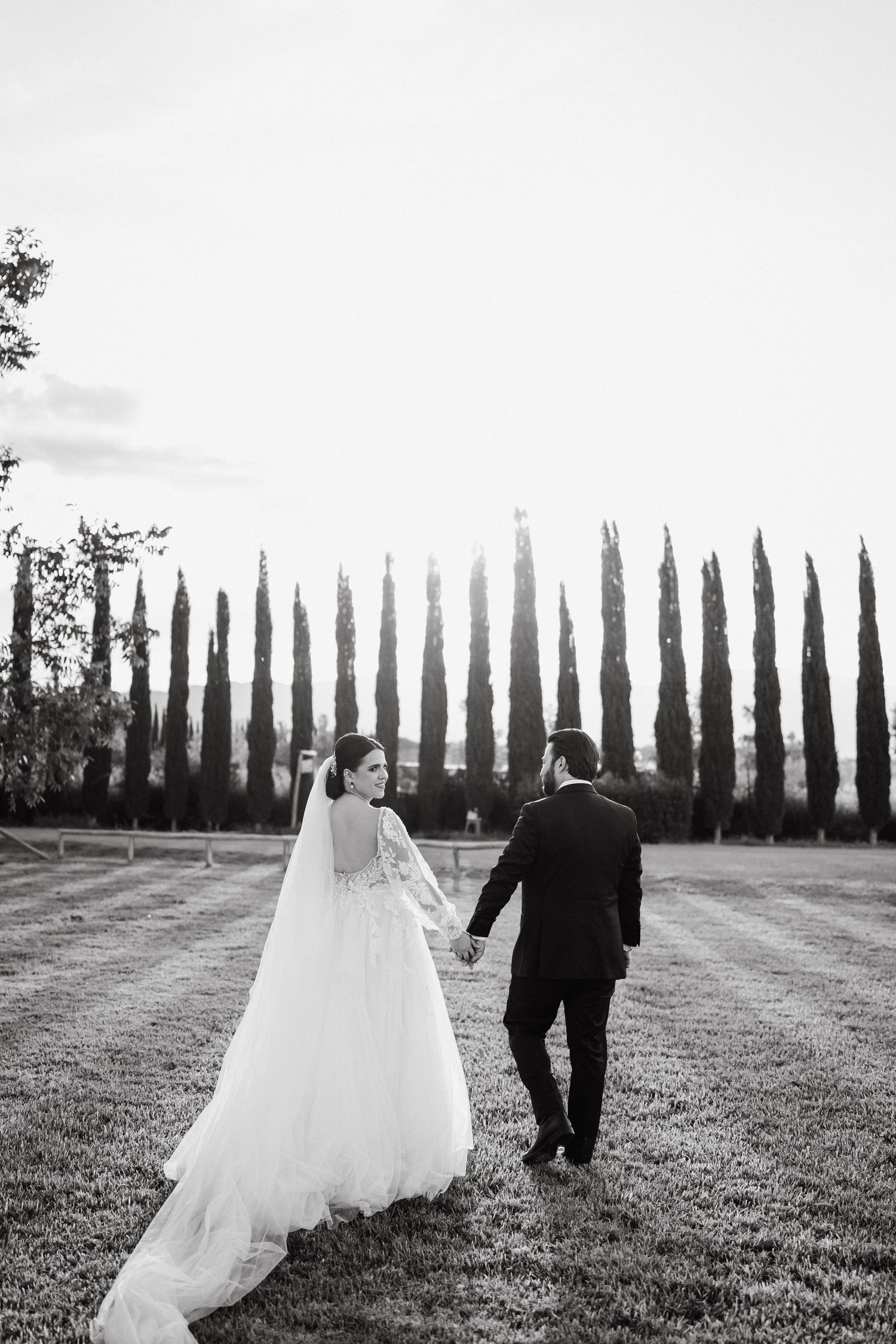 Black and white photo of bride and groom holding hands walking toward cypress trees on wedding day.