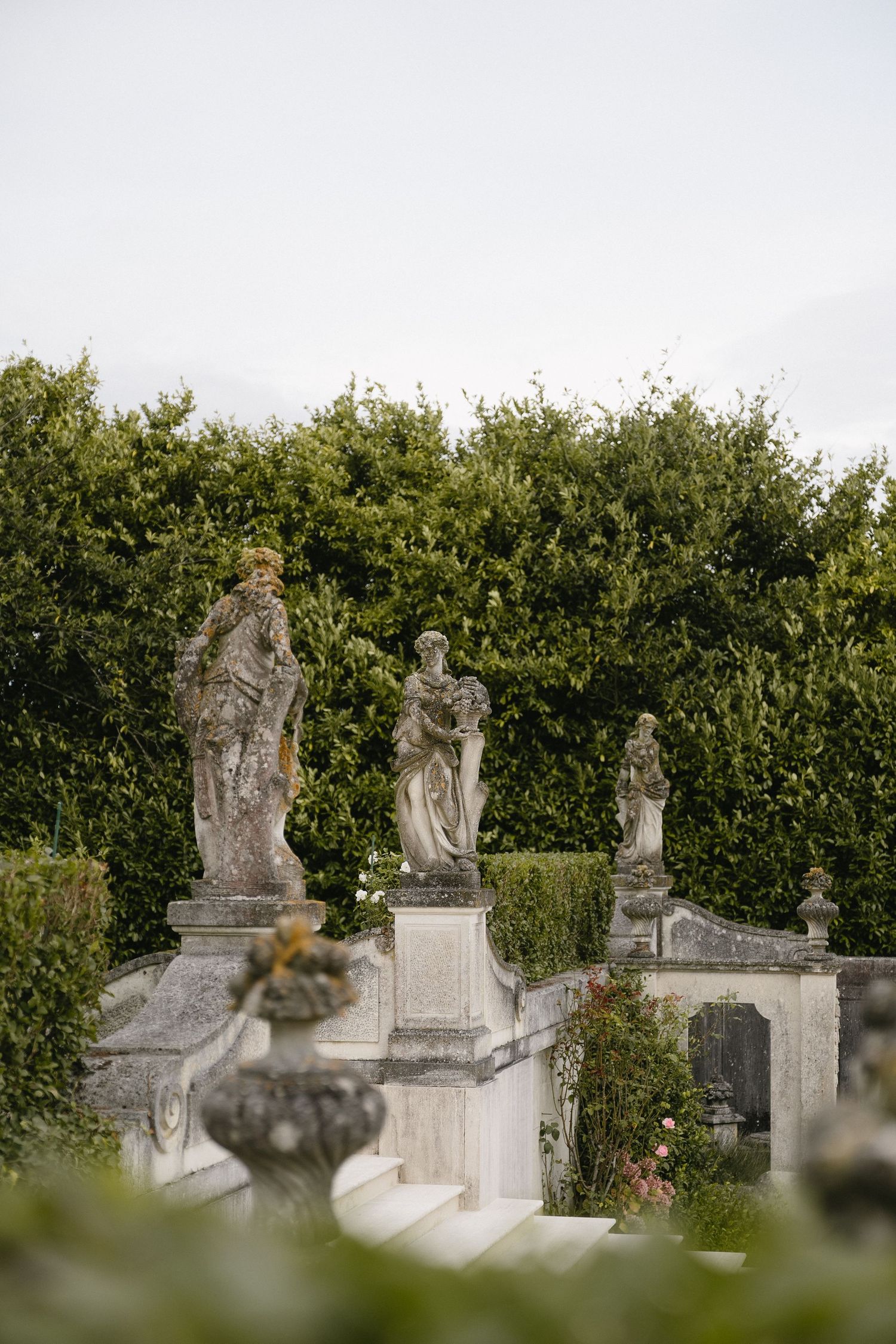 Stone angel statues and religious sculptures stand in a row along a garden wall with green hedges in the background.