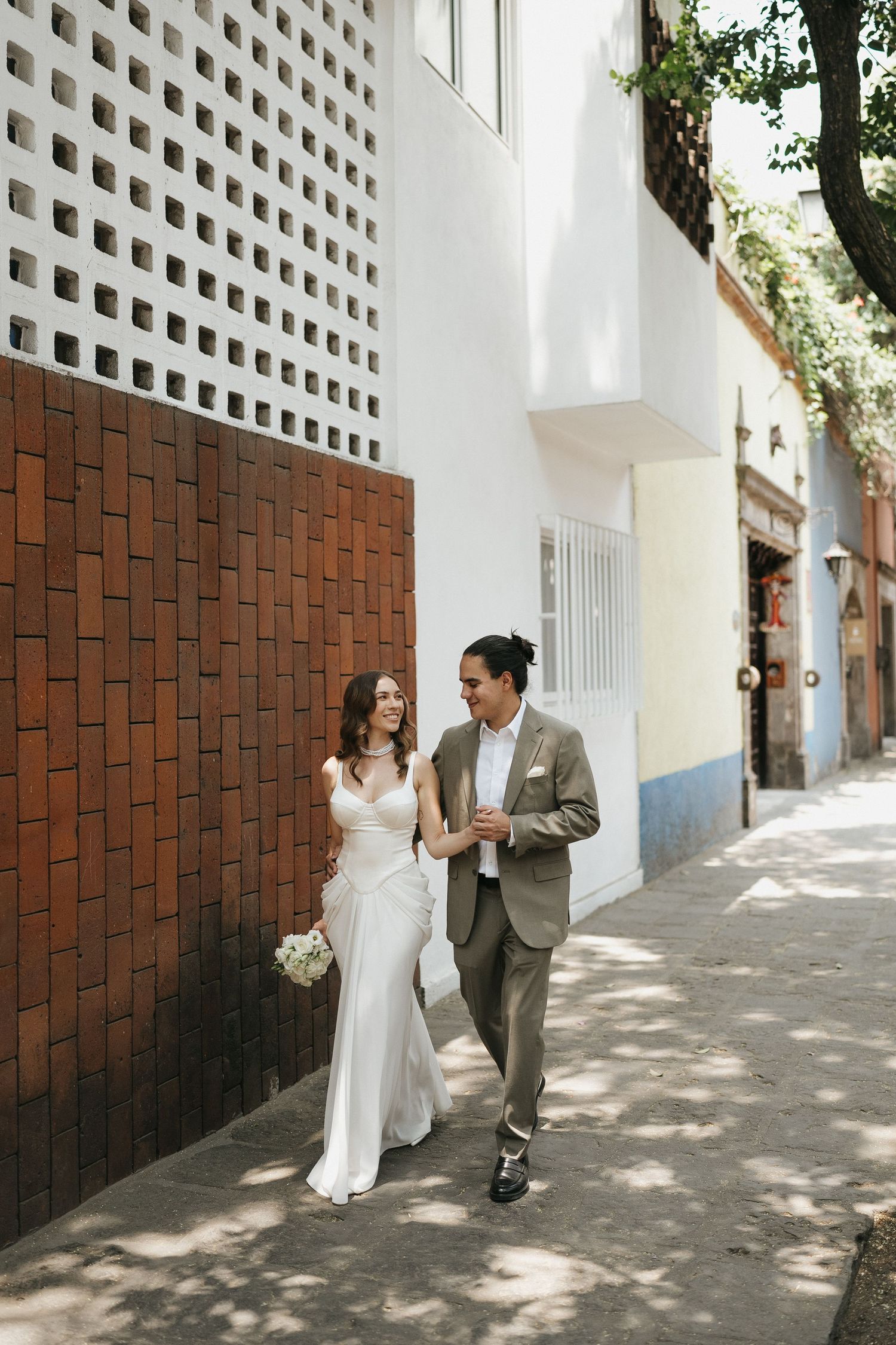 A couple in wedding attire walking along a brick wall with white lattice details.