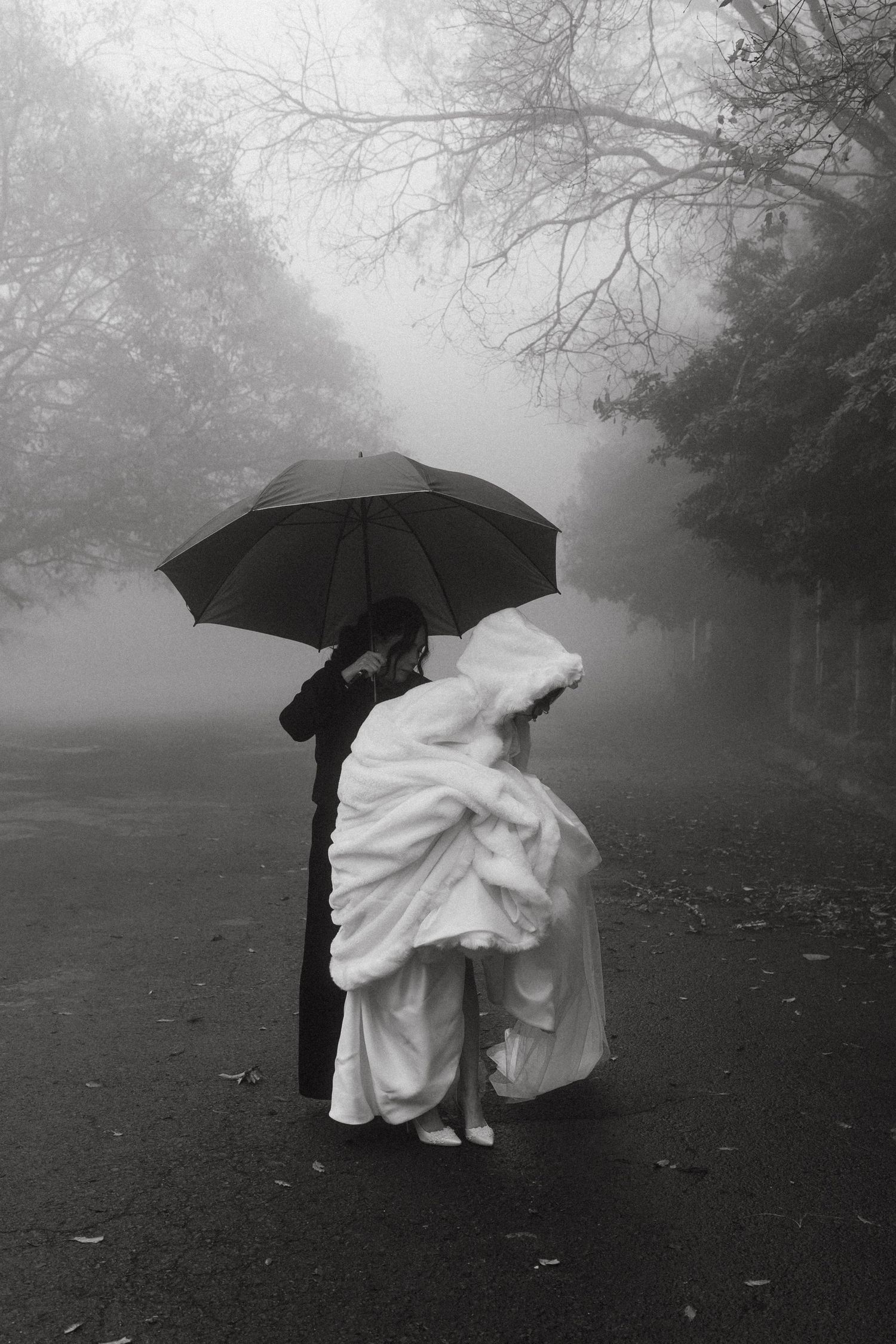 Ethereal scene of figures with umbrellas in foggy park wearing flowing white dresses.
