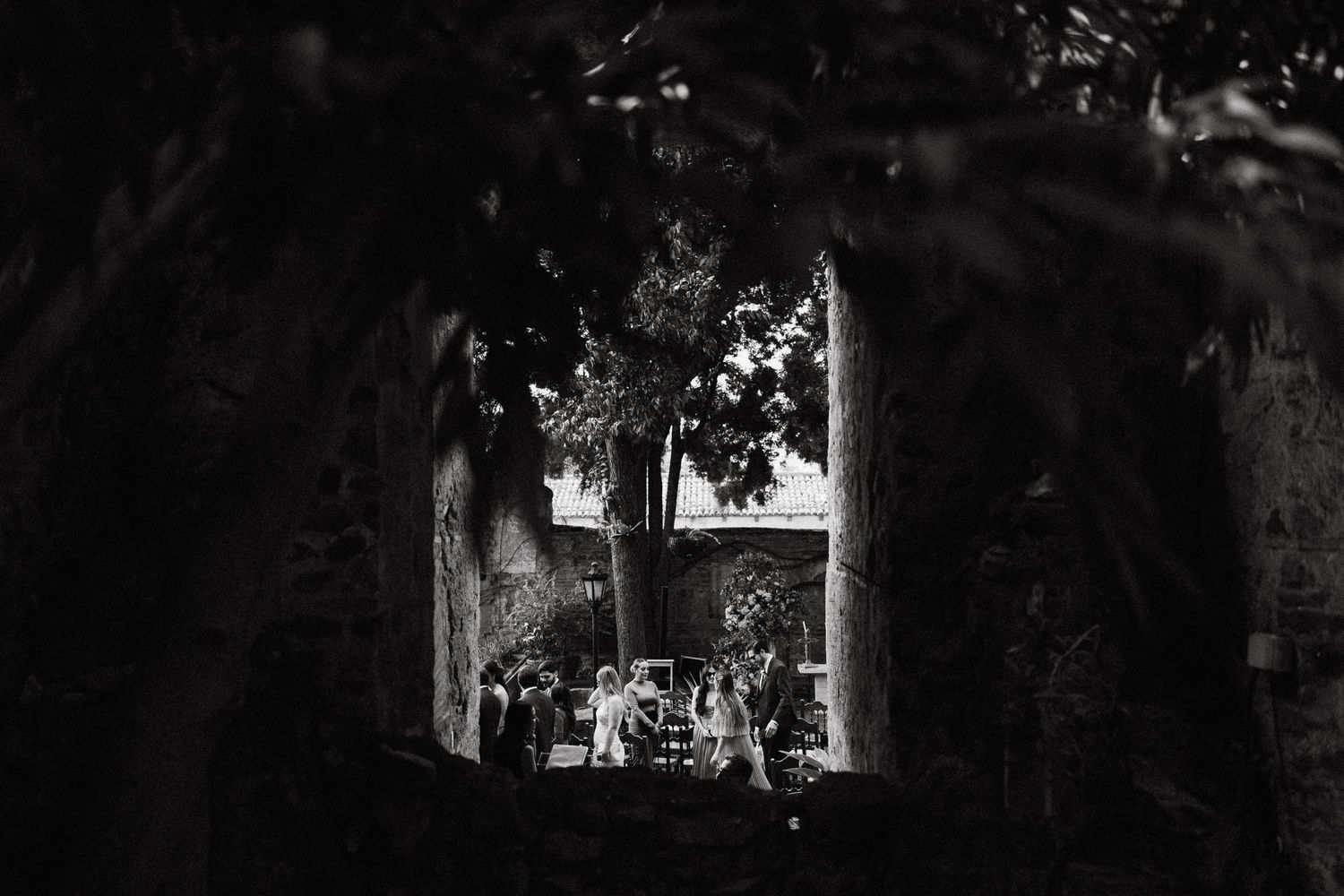 Black and white photo of people gathered in a sunlit courtyard framed by dark stone archway and lush trees.