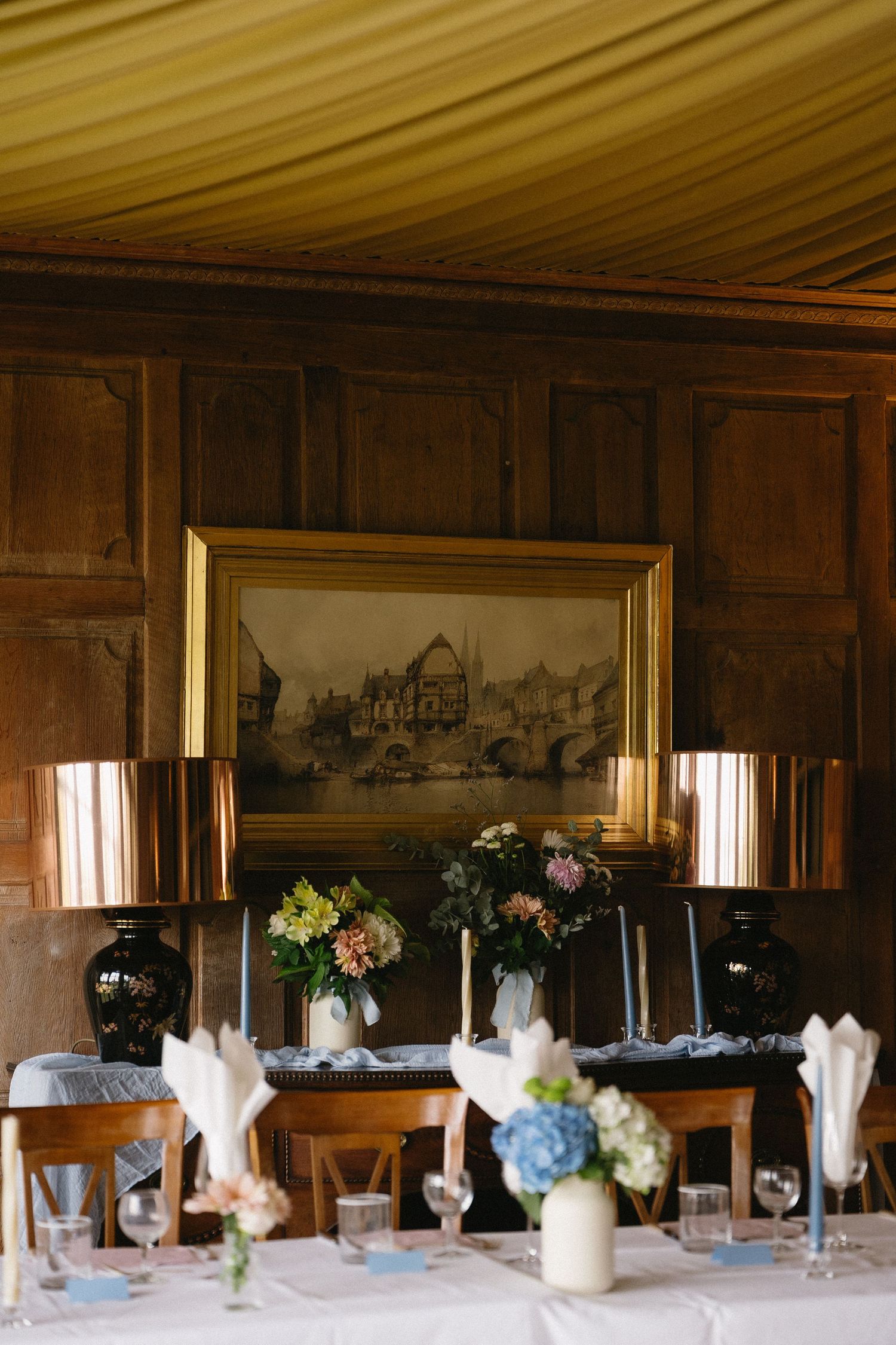 Elegant dining room table set for a wedding reception with white linens and floral centerpieces against wood-paneled walls.