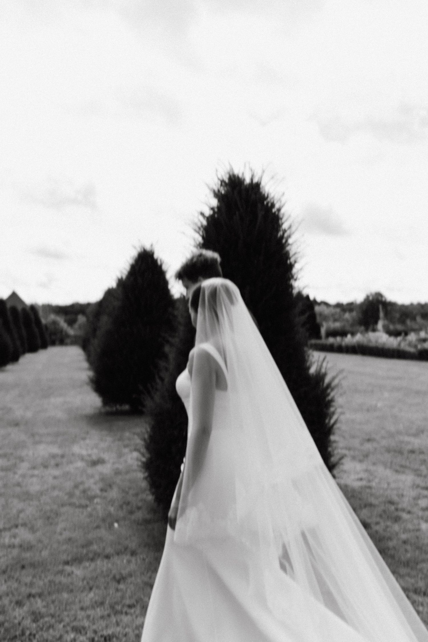 A black and white photo of a bride with flowing veil walking through a garden path lined with trees.