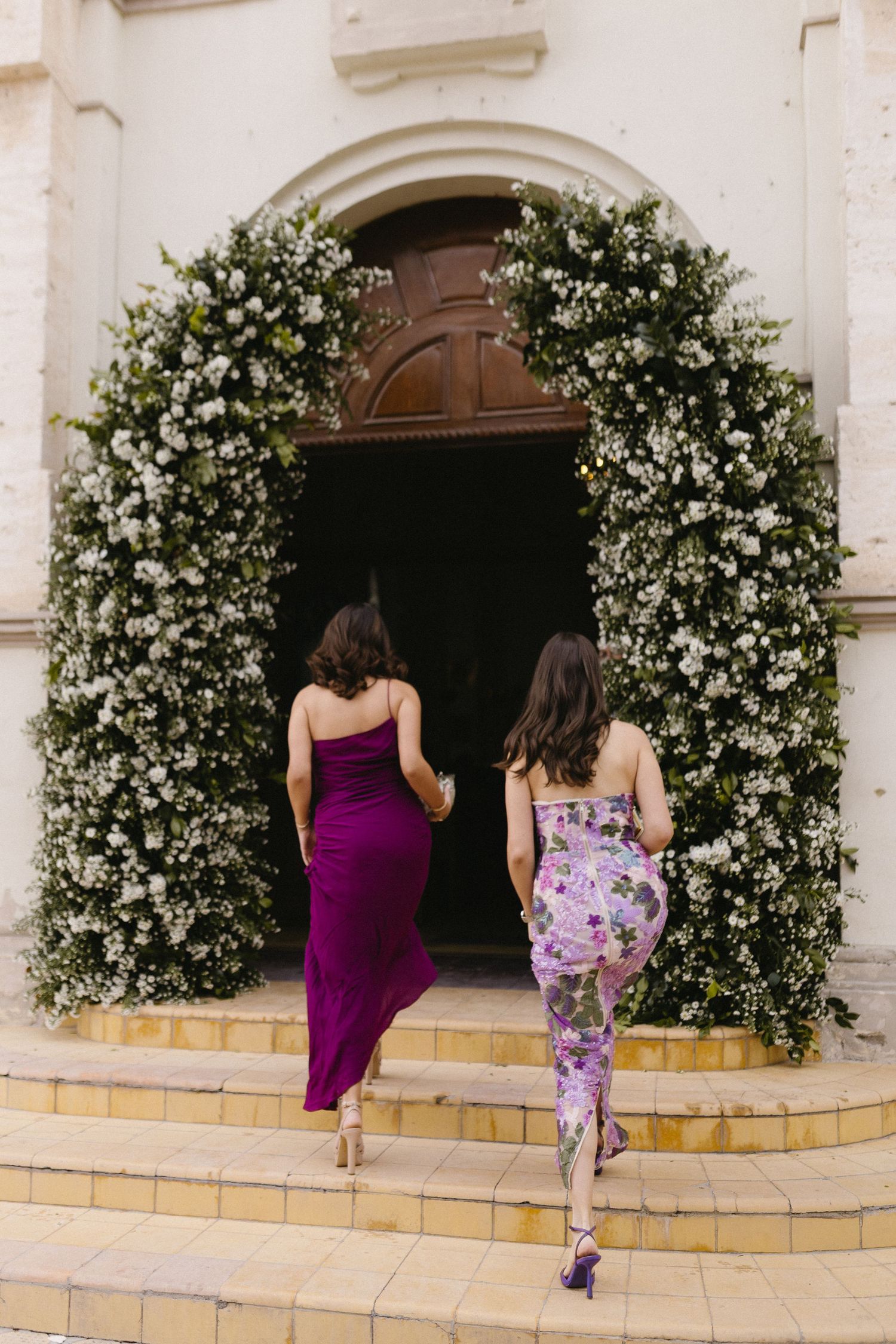 Bridesmaids in purple dresses walking past white floral archways outside a church.