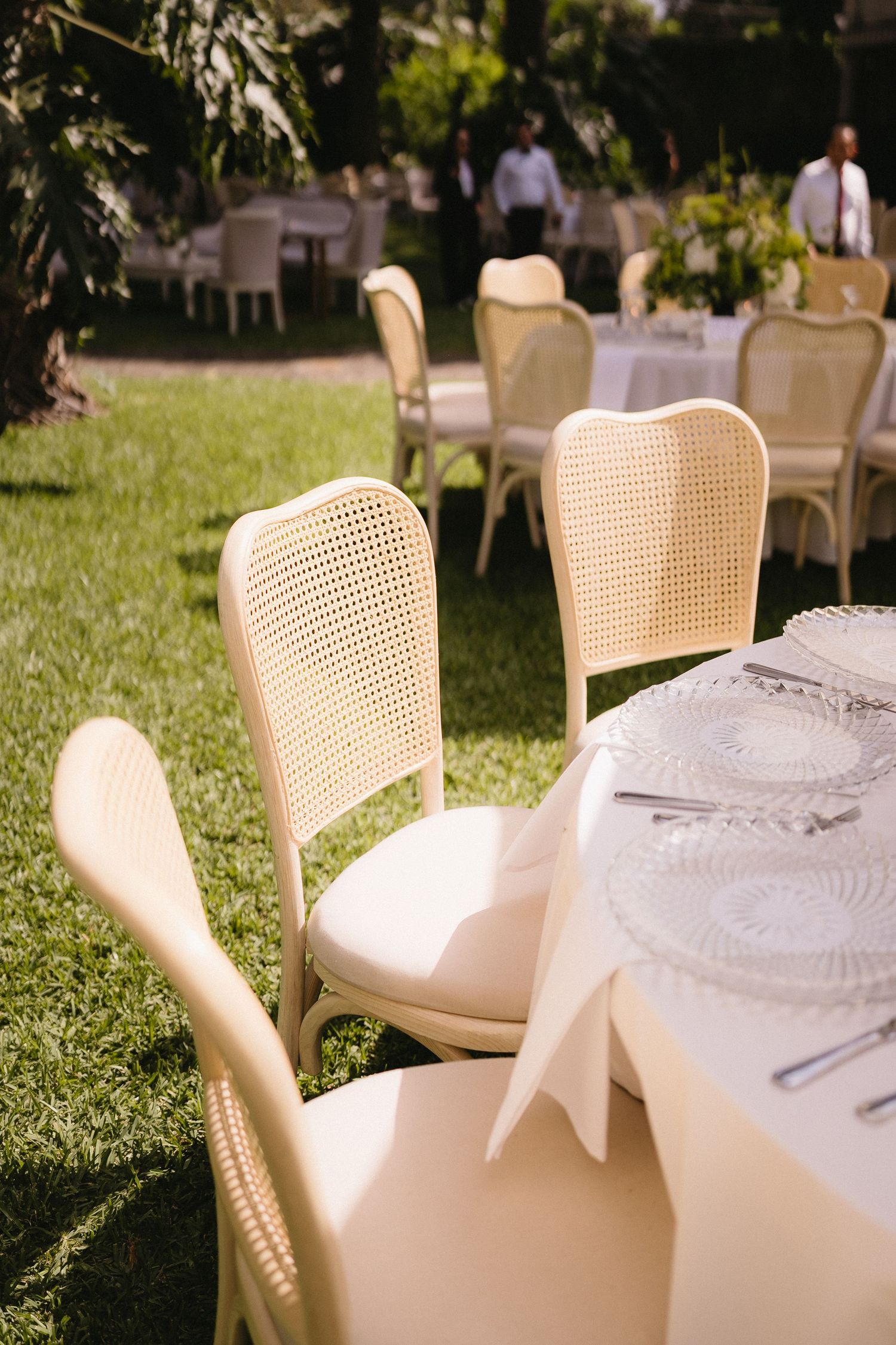 Elegant outdoor wedding reception setup with cane chairs and white table settings on grass.
