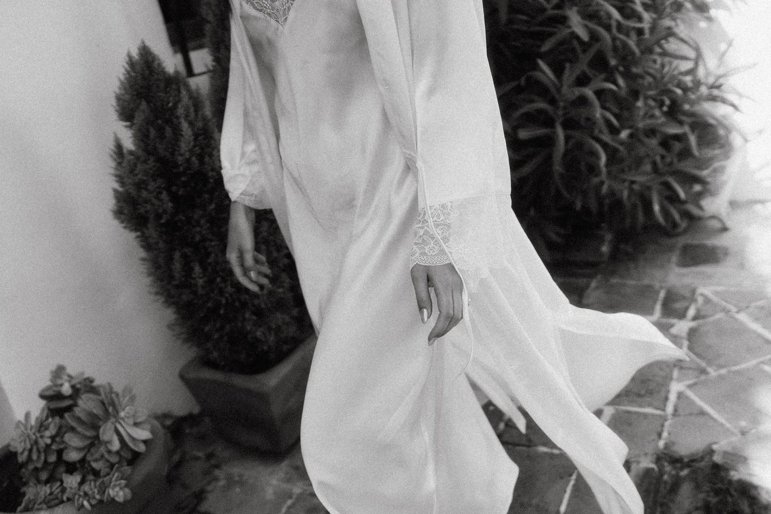 A bride in a flowing white wedding dress poses in an outdoor garden setting with lush greenery in the background.