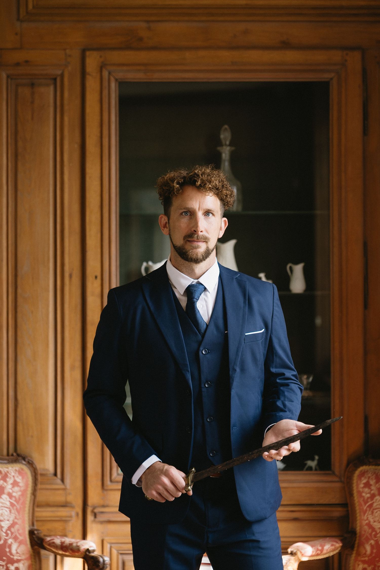 A groom in a tailored navy three-piece suit standing in front of wooden paneled walls.
