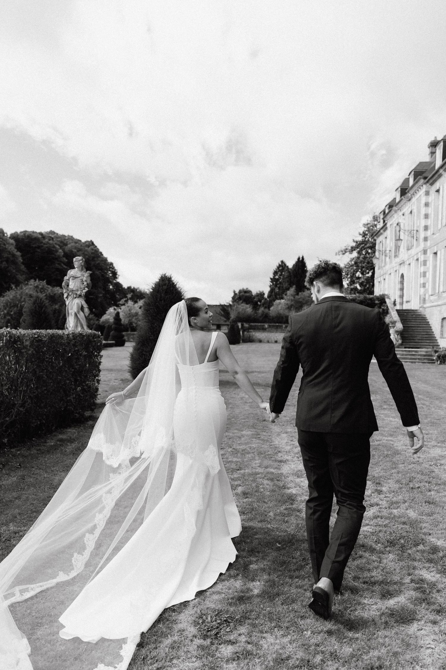 Wedding party walks across a grassy field with flowing veils trailing behind.