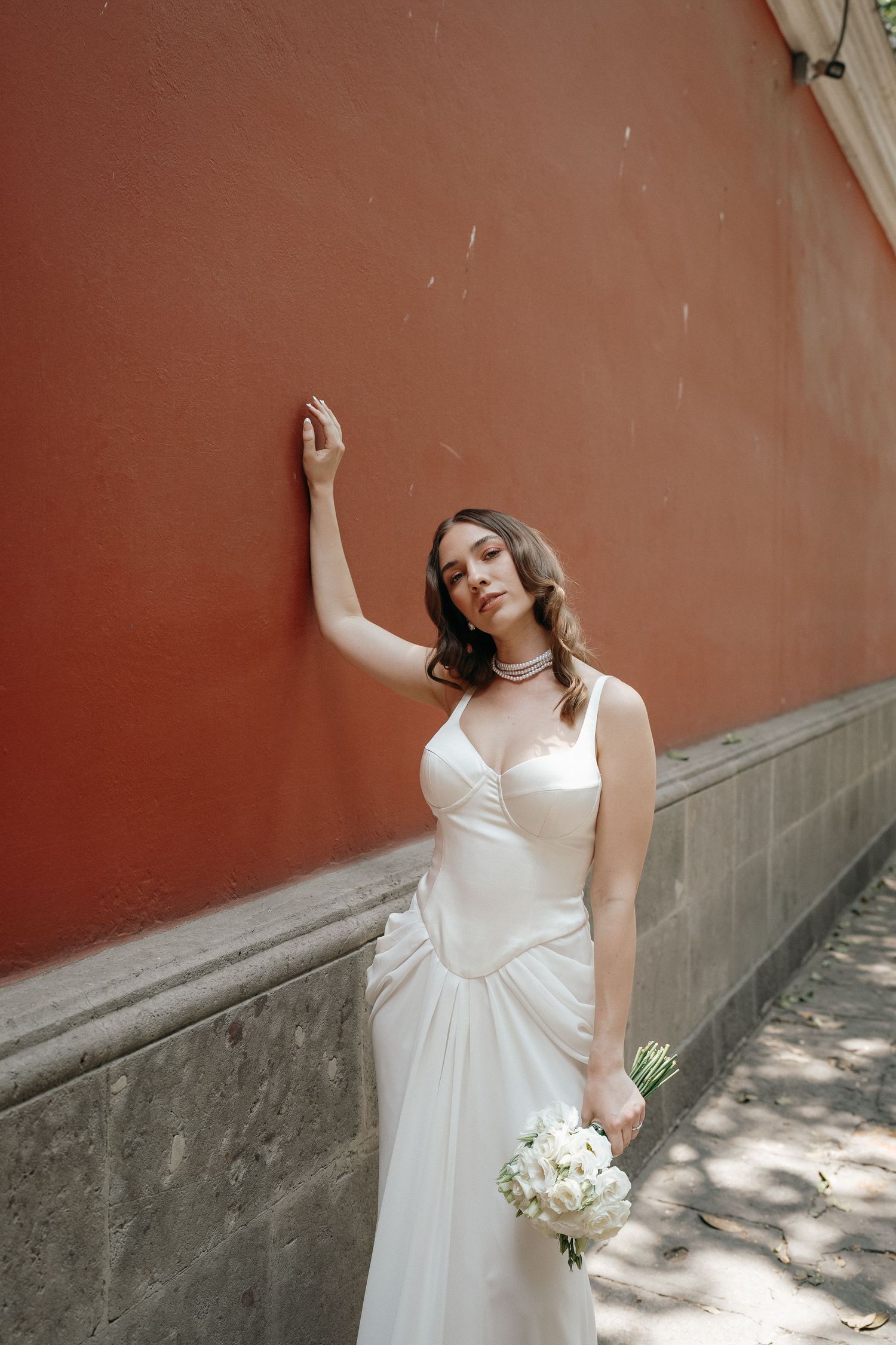 A bride in a white dress poses against a terracotta colored wall holding a bouquet.