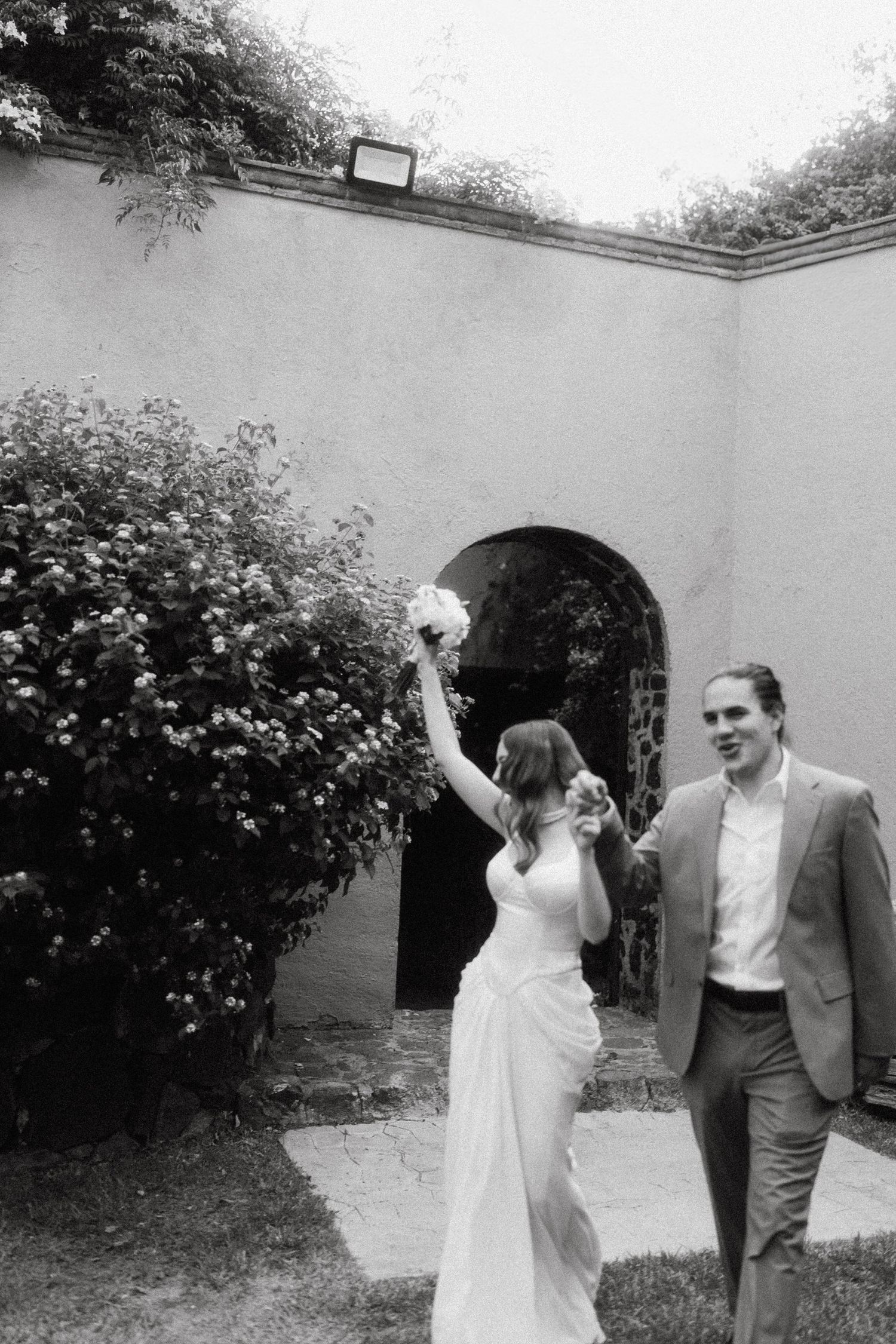 A joyful moment captured in black and white against a garden wall with flowering bushes.