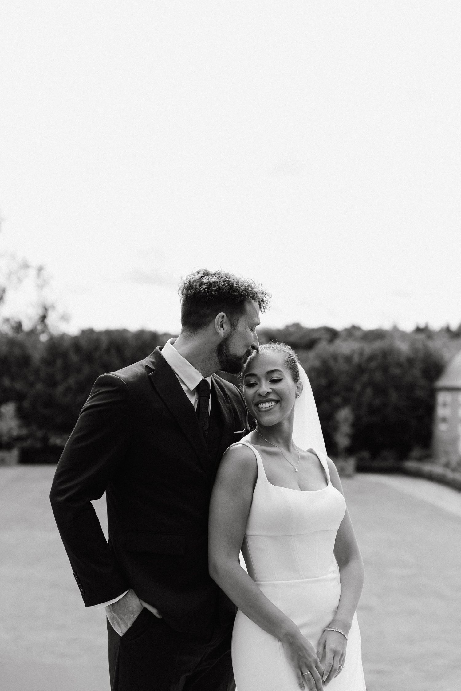 A couple shares an intimate moment in formal attire against an outdoor backdrop.
