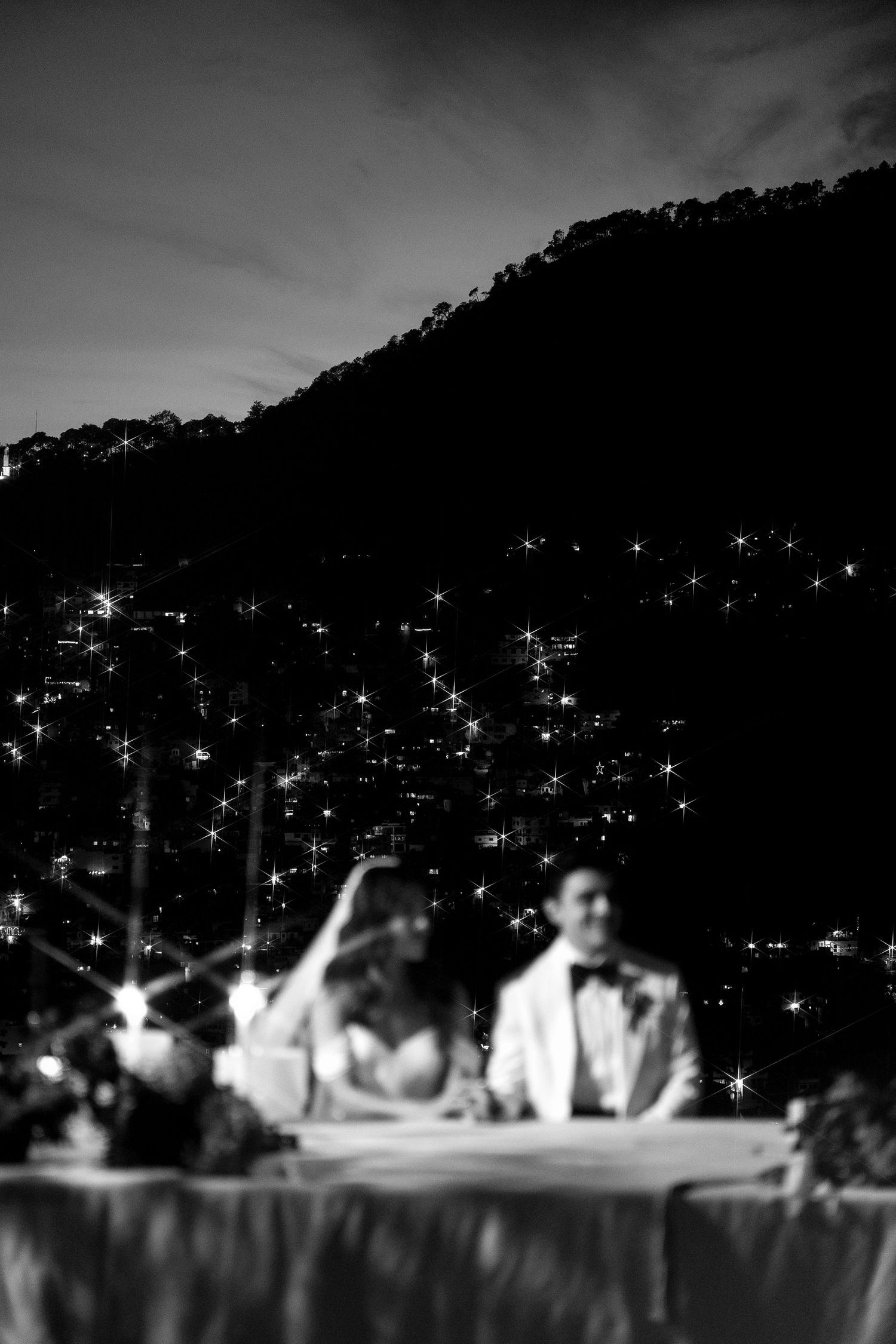A couple sits at a table during an evening outdoor reception with twinkling lights and a dark hillside in the background.