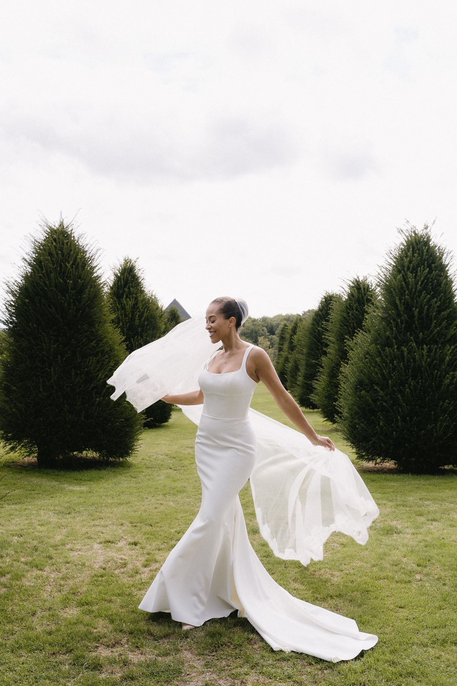 A bride in a white wedding dress twirls gracefully in a field surrounded by evergreen trees.