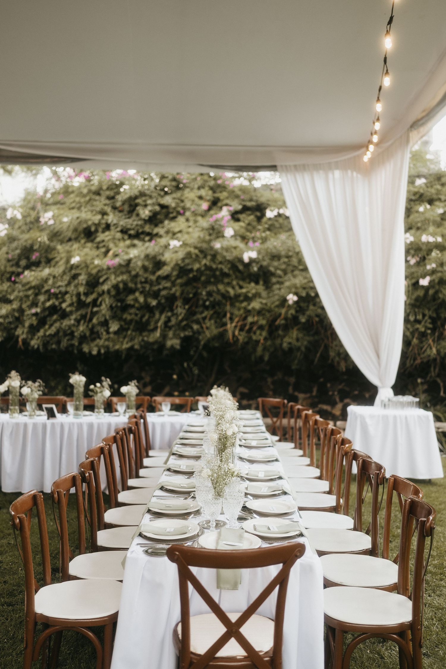Long banquet table with wooden chairs and white linens set for an outdoor wedding reception.