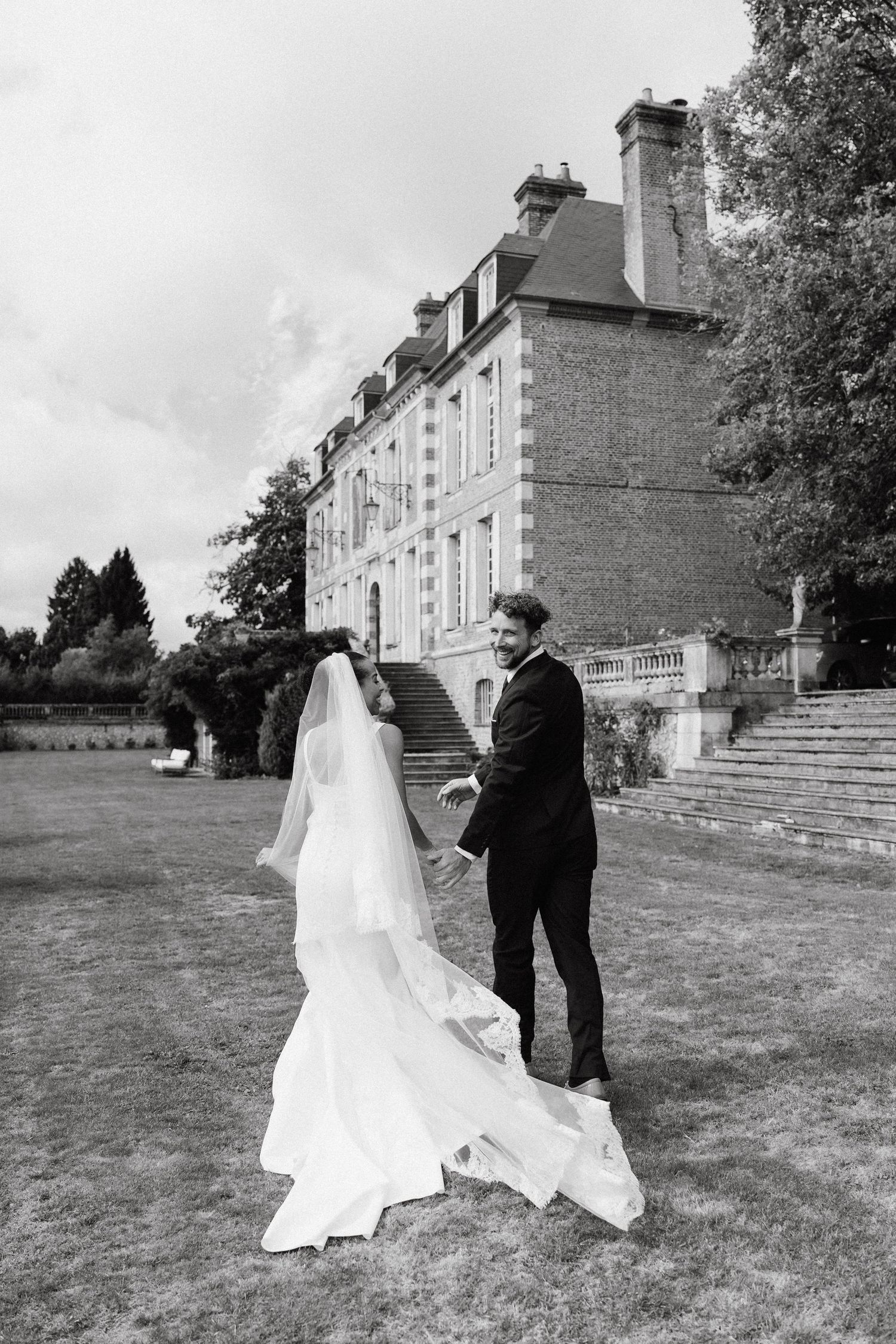 A black and white wedding photo of a couple standing on the lawn of a grand French chateau with stone architecture.
