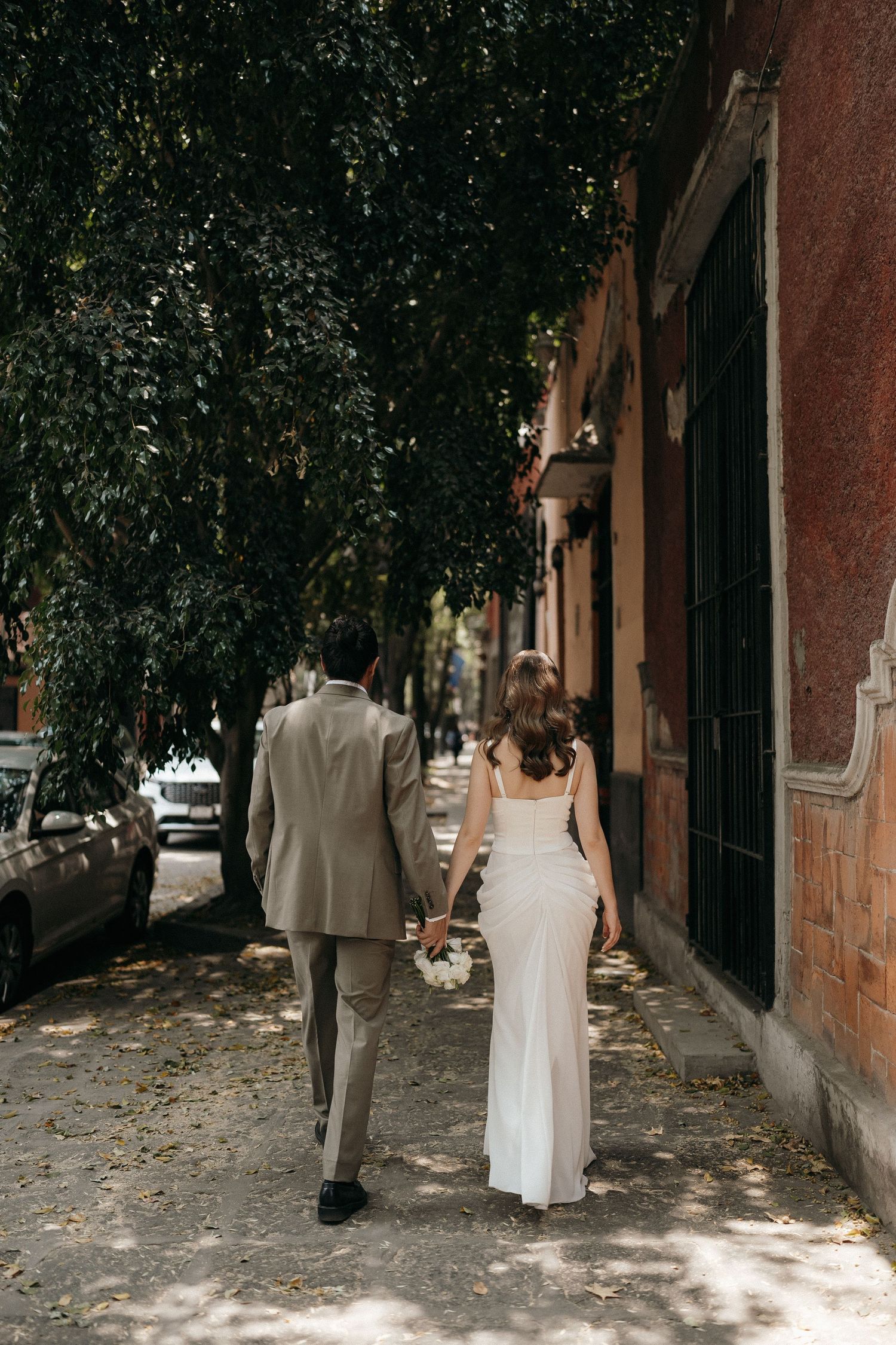 Wedding couple walking through a tree-lined urban pathway.