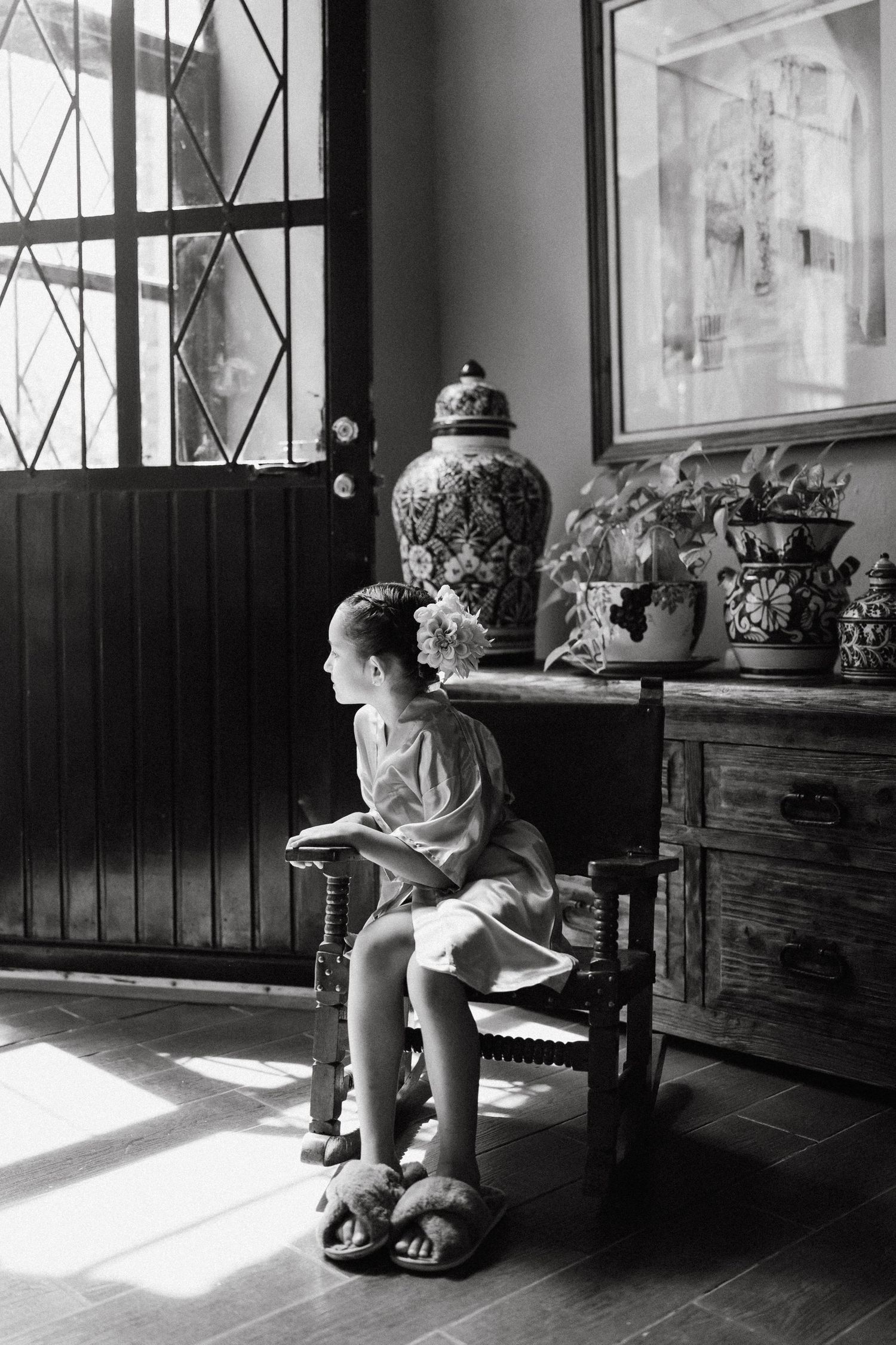 Black and white photo of a child sitting in a wooden chair by a window with dramatic lighting.
