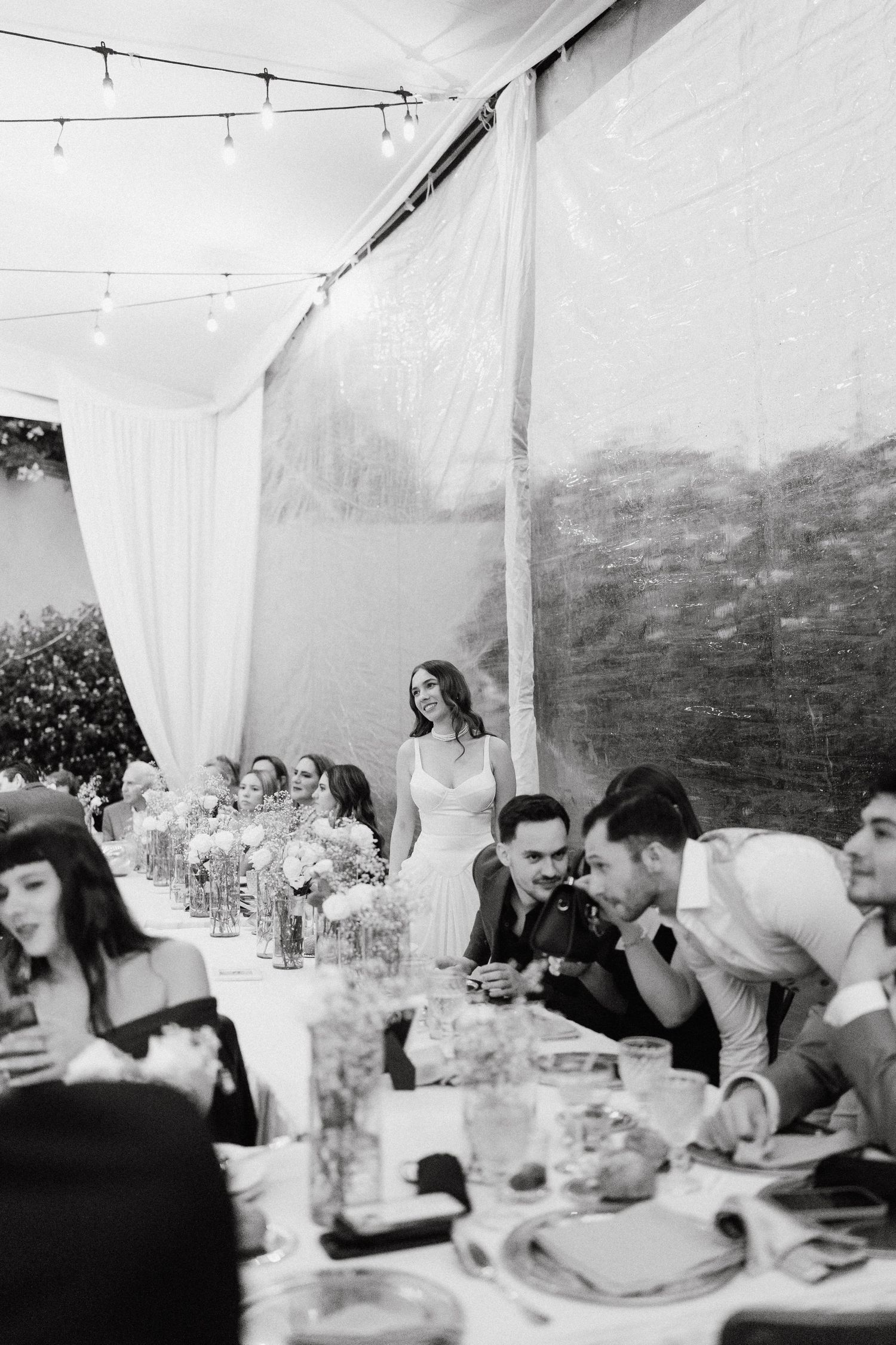 Black and white photo of wedding reception tables under white tent with string lights.