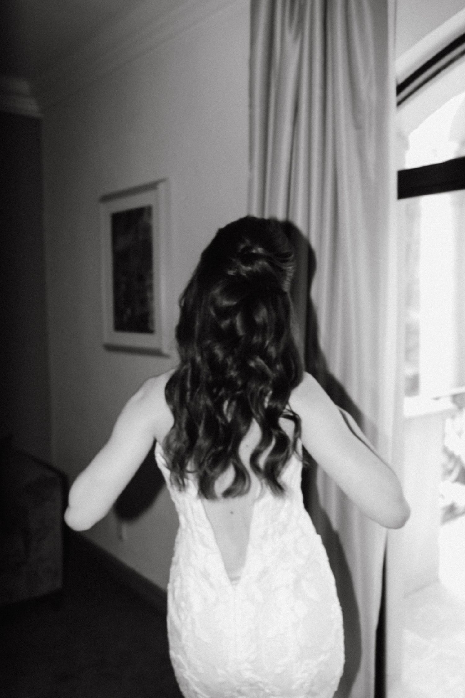 Back view of a white lace wedding dress being adjusted in natural window light.