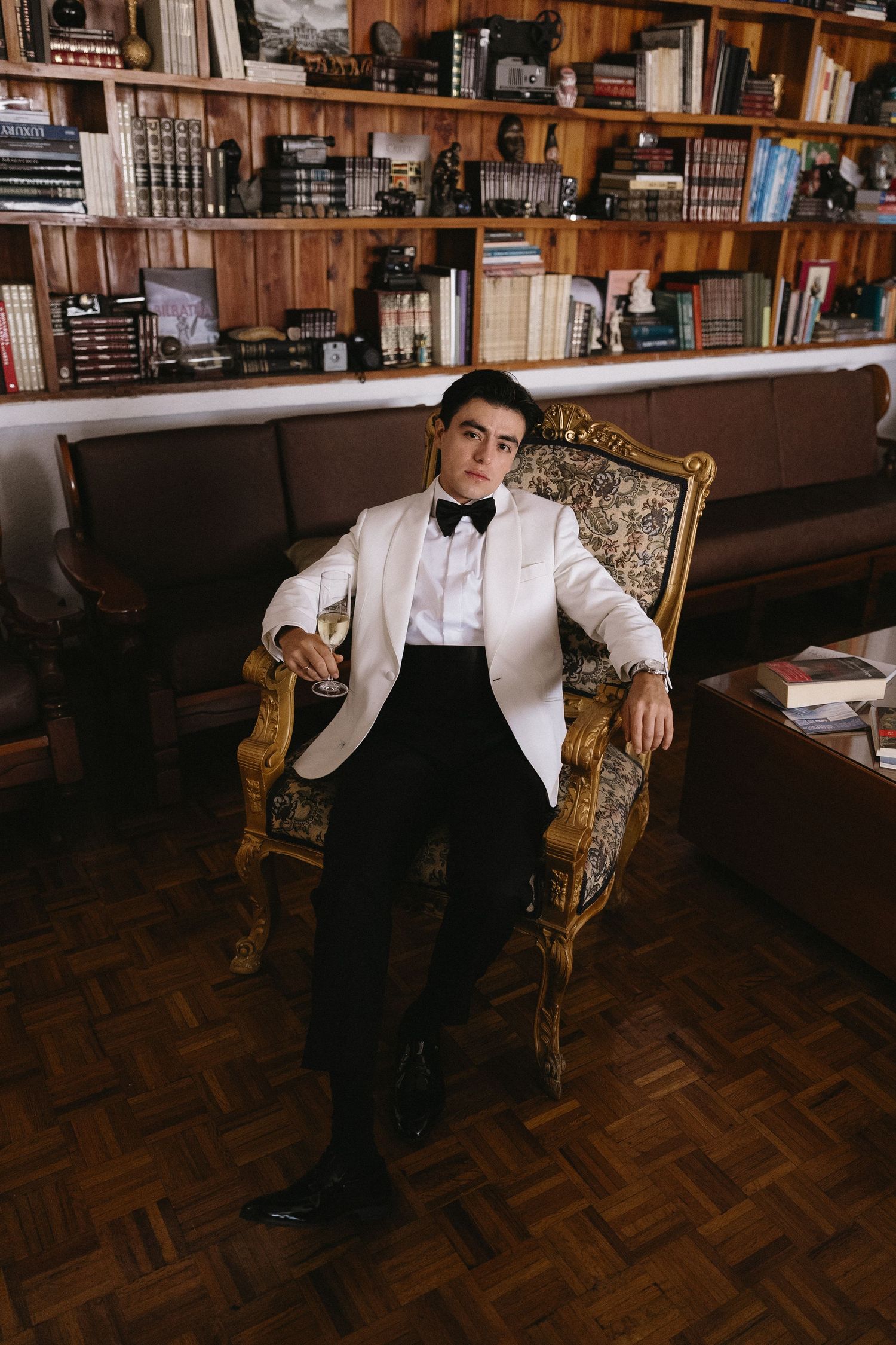 A person in formal attire sits on an ornate gold chair in a library with wooden floors and bookshelves.