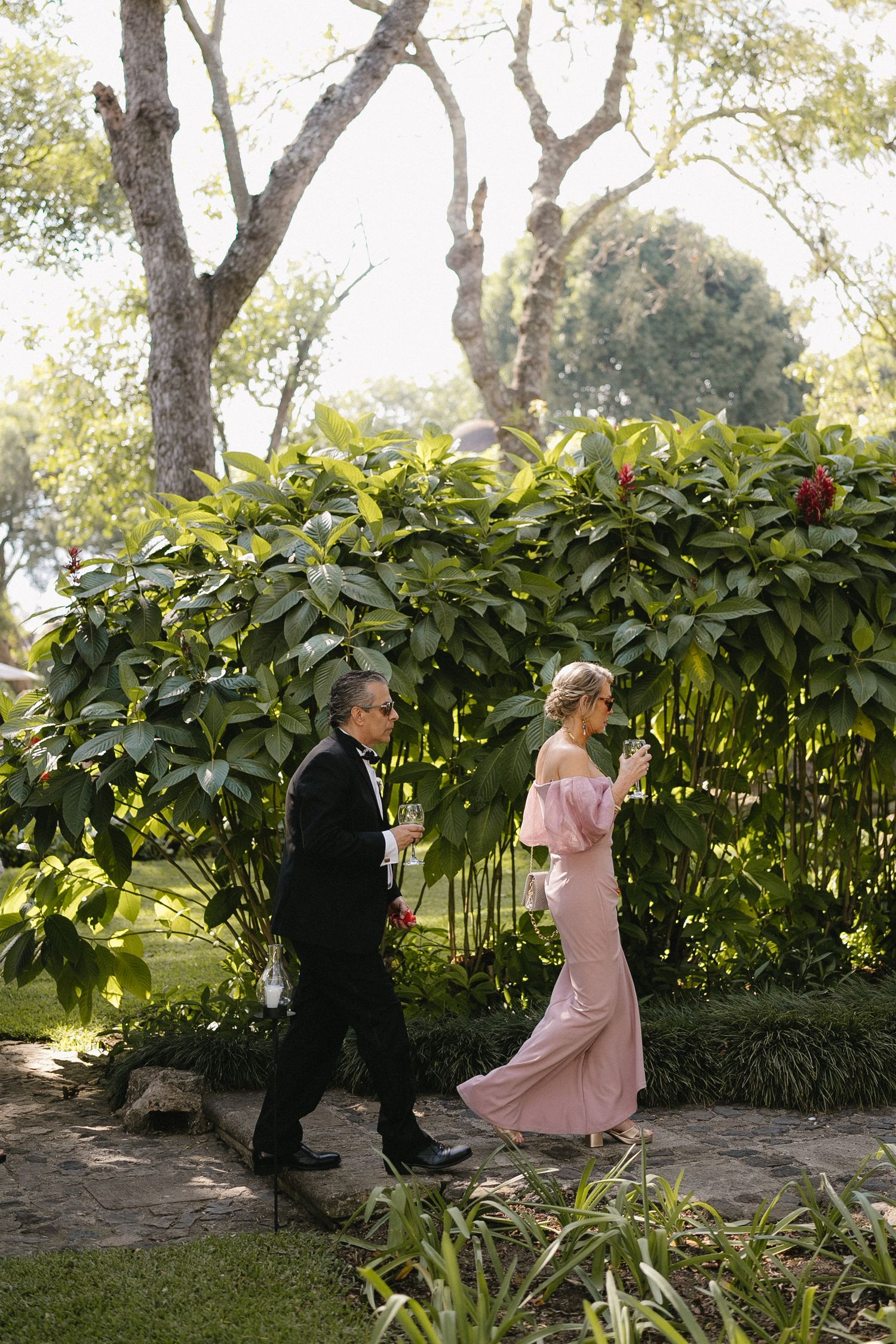 Wedding party walking through garden path with lush greenery and trees.