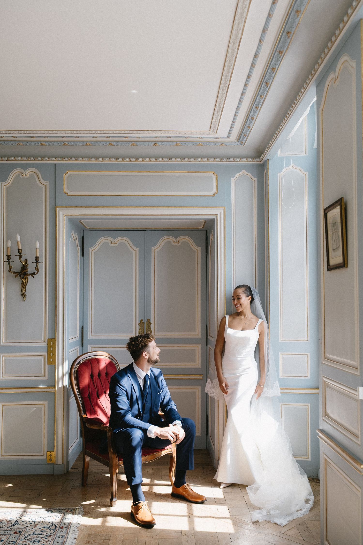 A wedding couple poses in an elegant blue-walled room with ornate moldings and natural light.