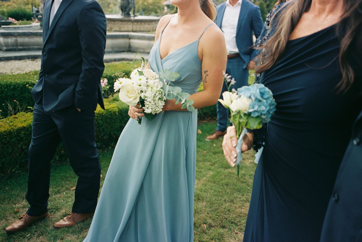 Wedding party members in light blue bridesmaid dresses and navy suits gather outdoors for formal photos.