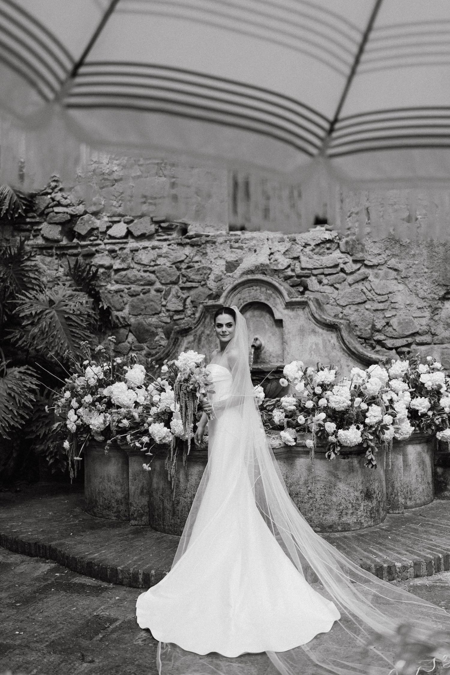 Black and white photo of a wedding dress with flowing train against a stone wall backdrop.